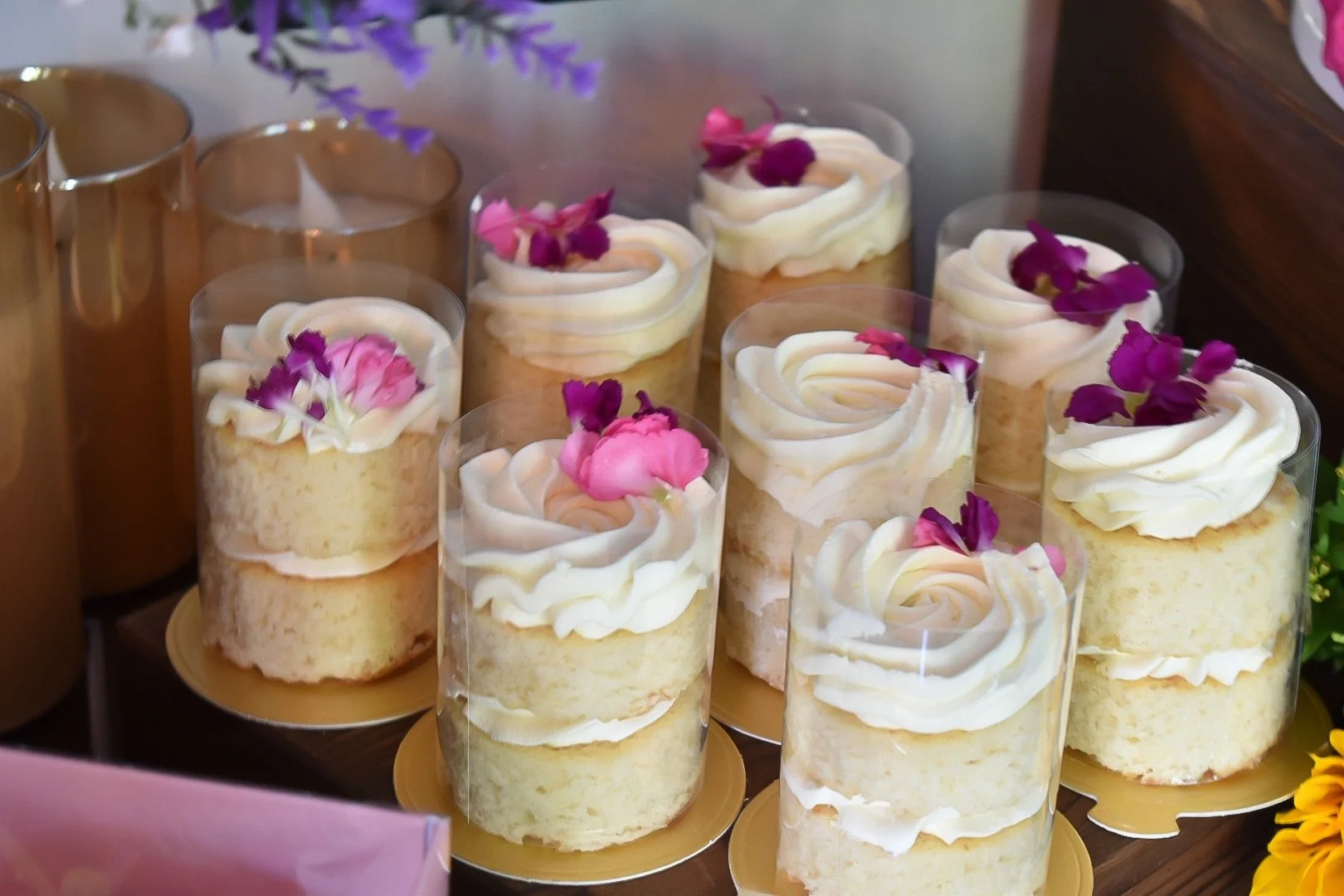 A collection of vanilla sponge cake desserts topped with white whipped cream, pink and purple flower petals, served in tall clear glasses on golden trays.