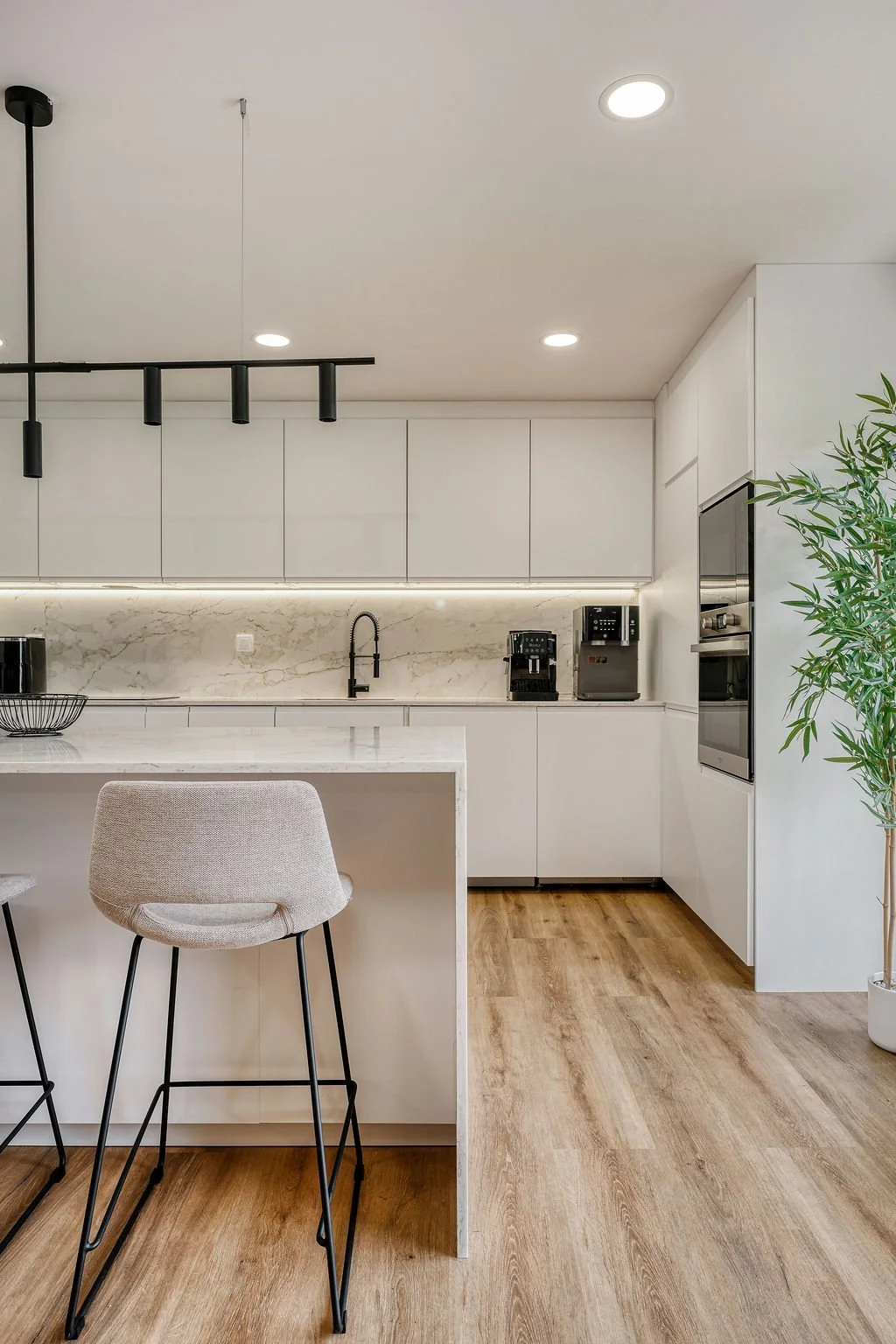 Cozinha moderna com armários brancos, bancada de mármore, eletrodomésticos pretos e um balcão com duas banquetas e uma planta na esquina