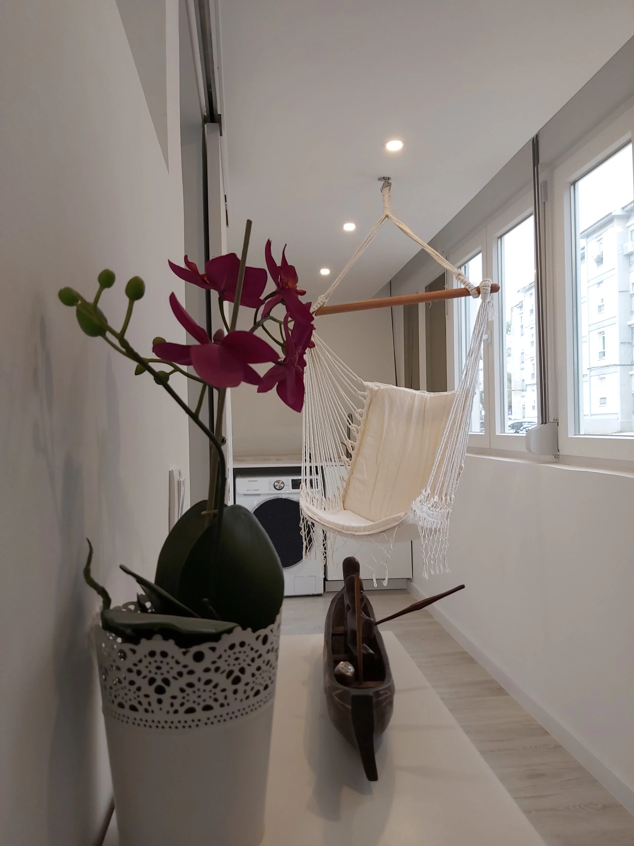 Vaso com orquídea roxa na esquerda e uma colher de pau na altura da flor, uma vela na cor marrom no centro, uma rede branca de descanso pendurada do teto ao lado da janela grande, dentro de um apartamento com paredes brancas e piso de madeira clara.