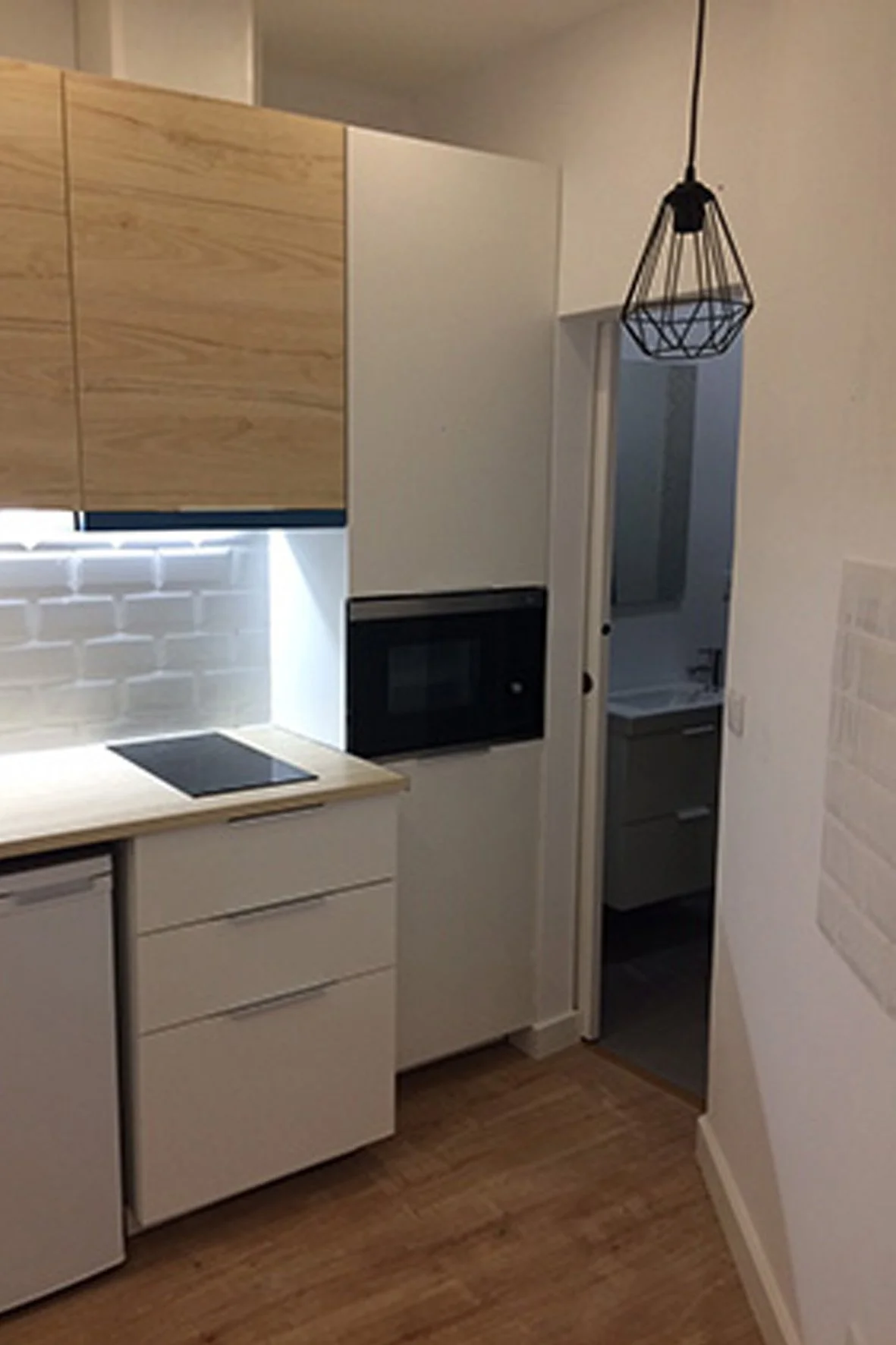 Cozinha compacta com armários brancos, cooktop, micro-ondas embutido, paredes de tijolos brancos, piso de madeira e luminária decorativa preta.
