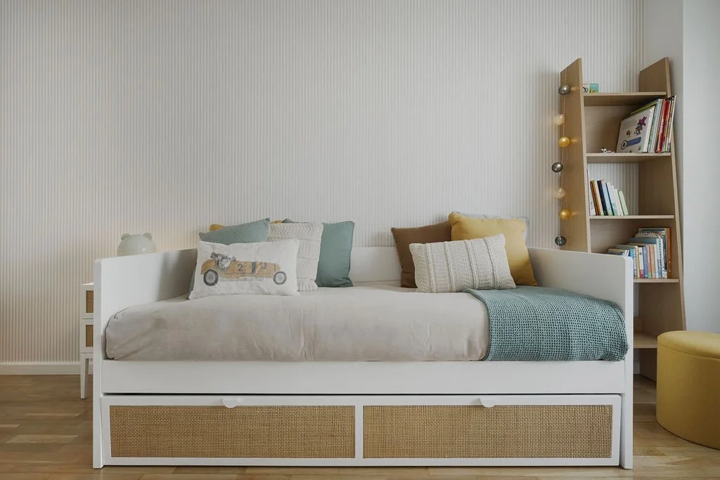 Quarto com cama de solteiro com almofadas, livros na estante de madeira inclinada, parede de fundo com textura de linhas, piso de madeira, cadeira amarela ao lado.