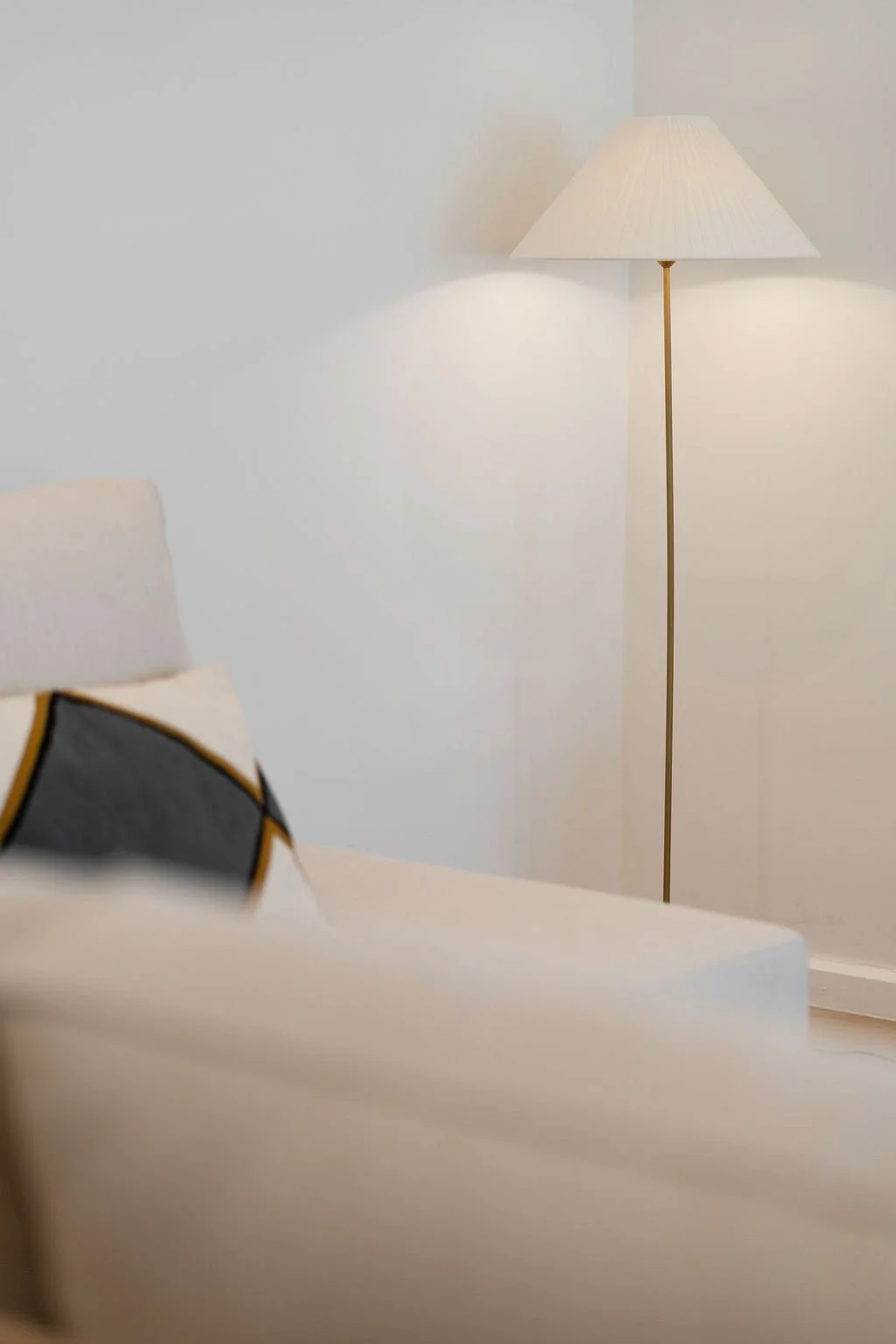 Sala de estar com parede branca, lâmpada de pé com abajur branco e sofá branco com almofada decorativa cinza e preta com detalhes dourados.
