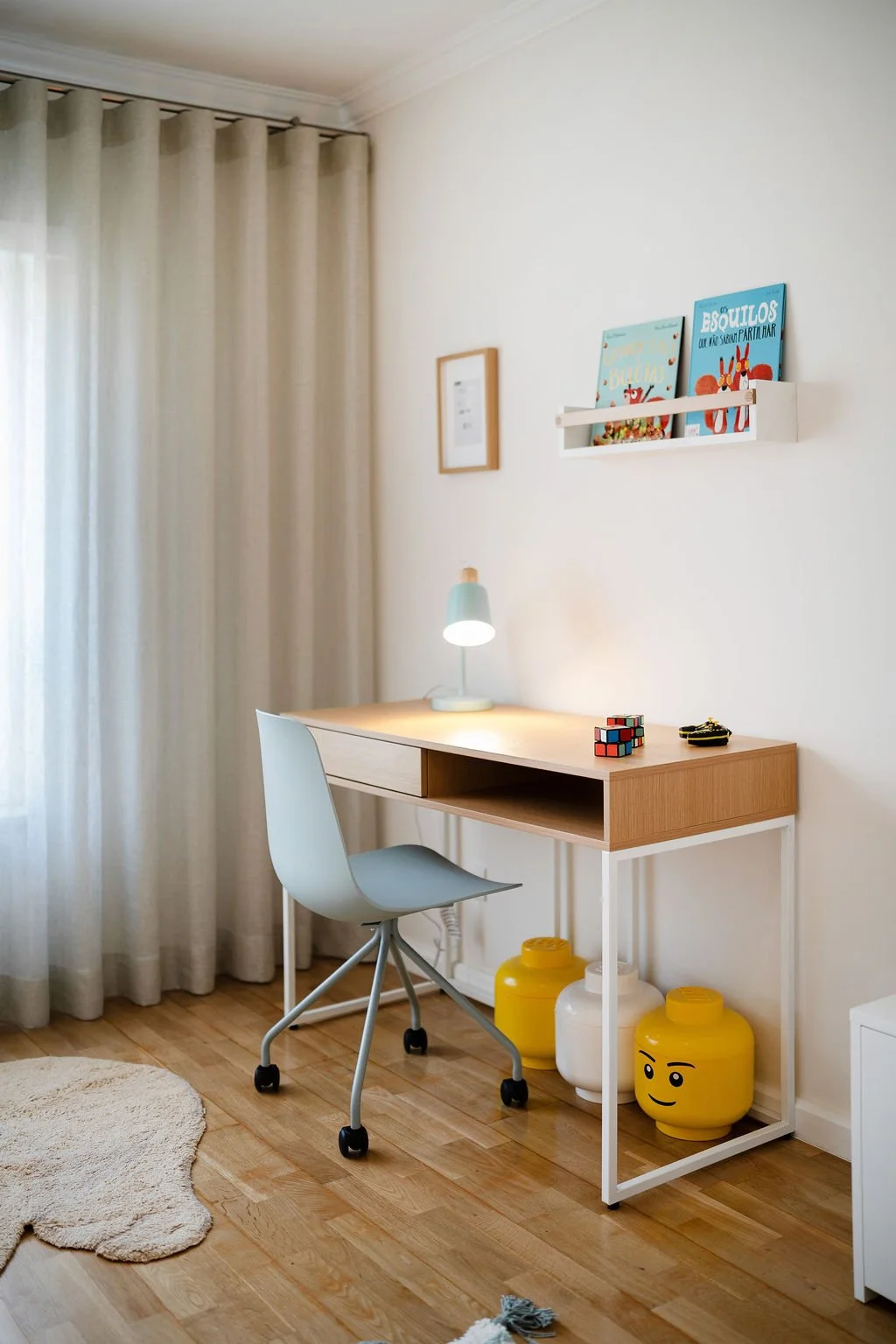 Escrivaninha de estudo com luminária, cubo Rubik e objetos decorativos, cadeira cinza, parede com quadros e prateleira com livros infantis, cortina branca de tecido ao lado de janela, chão de madeira e tapete bege ao lado.