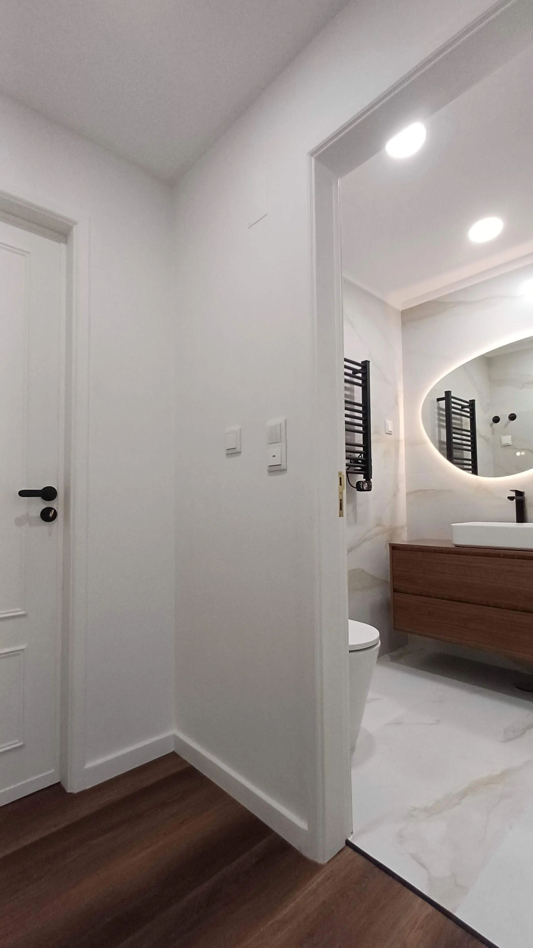 Banheiro moderno com espelho com iluminação de led, pia de bancada de madeira, vaso sanitário, aquecedor de toalhas preto e paredes de mármore branco com veios cinza.