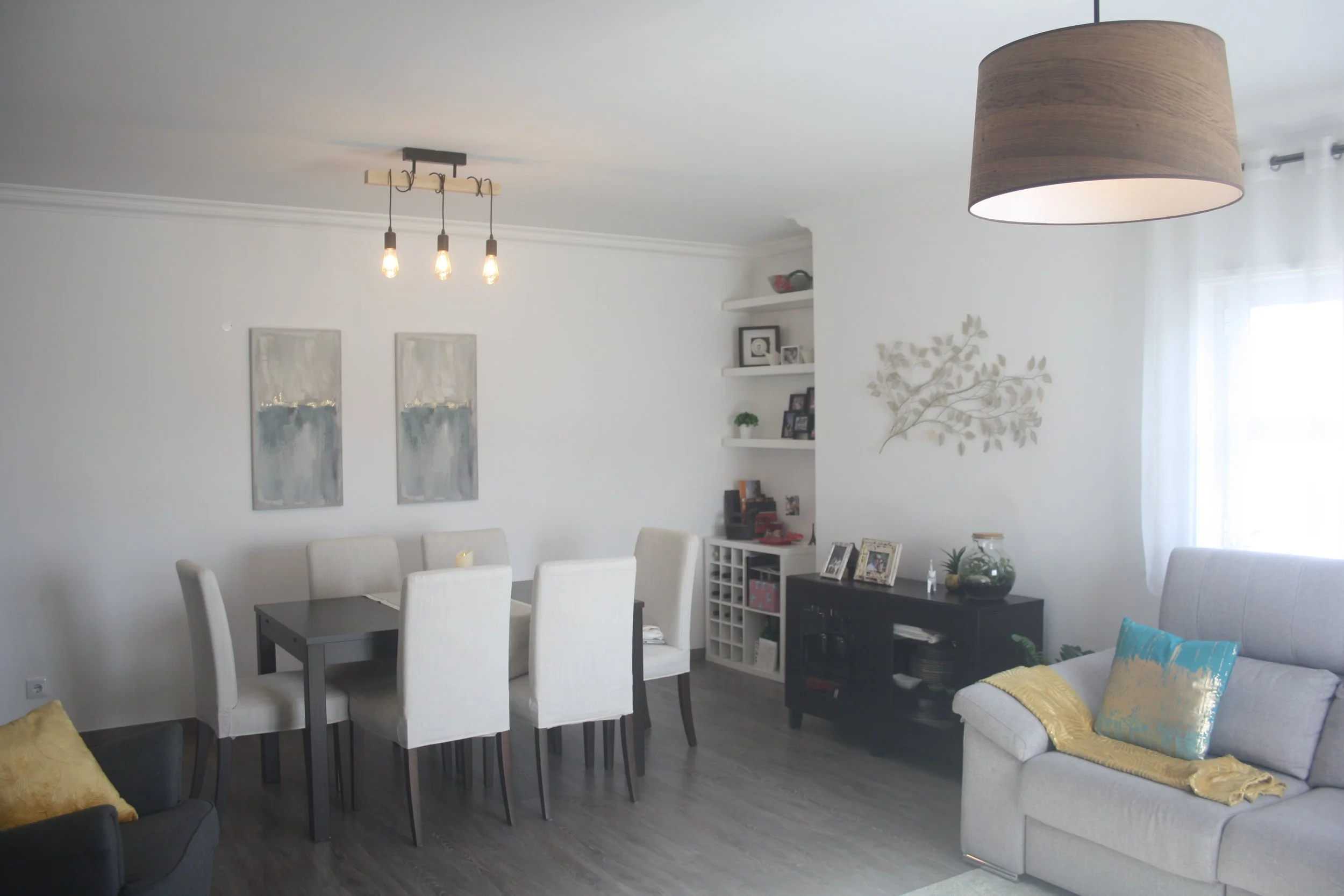 Sala de estar com mesa de jantar, cadeiras brancas, sofá cinza, luminária de teto de madeira, parede branca com quadros, prateleiras com fotos e objetos decorativos, cortinas brancas, piso de madeira escura.