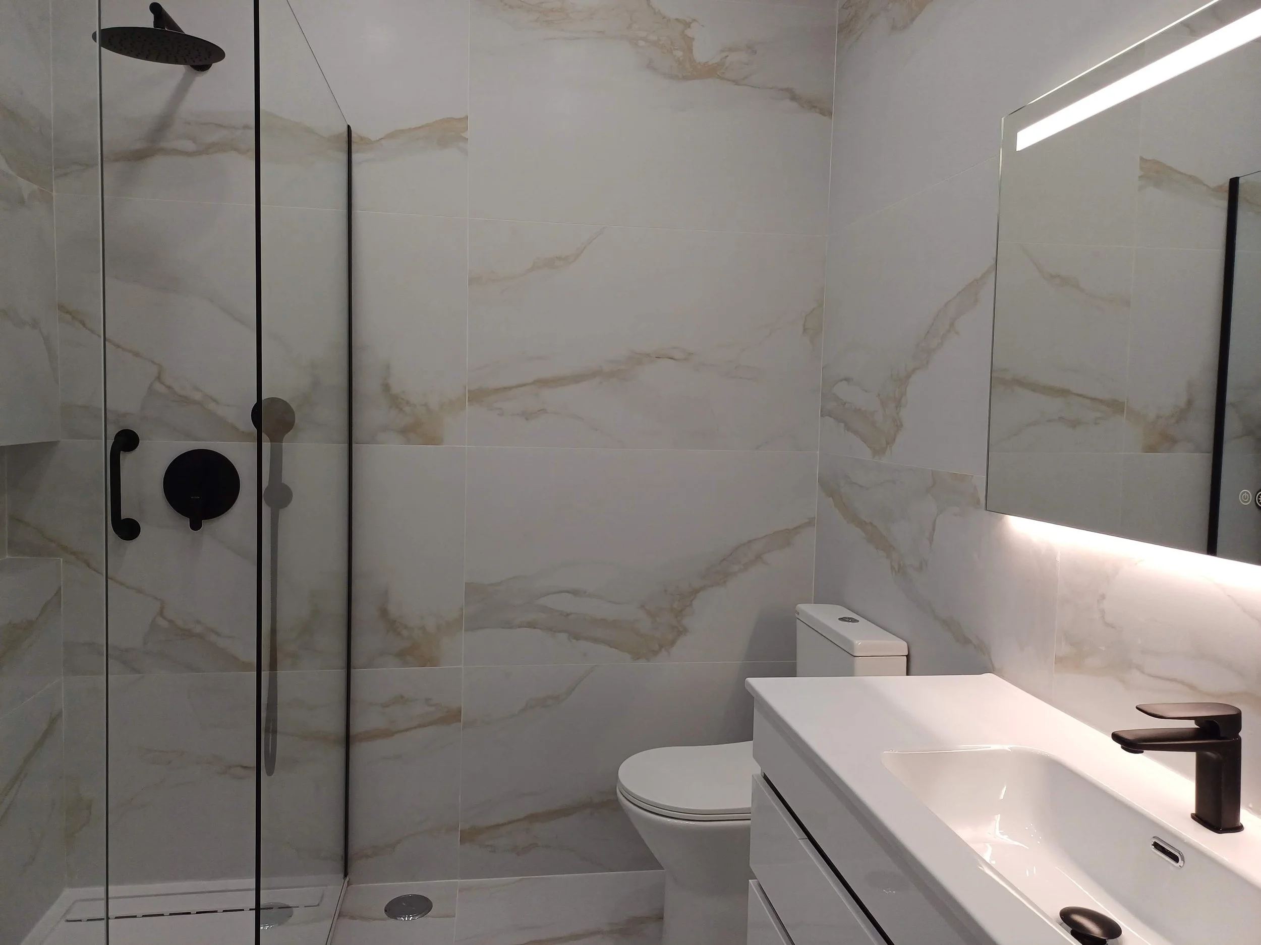 Banheiro com paredes de mármore, ducha com vidro de correr, banheiro com vaso branco e pia branca de balcão com torneira preta, espelho retangular com iluminação ao redor.