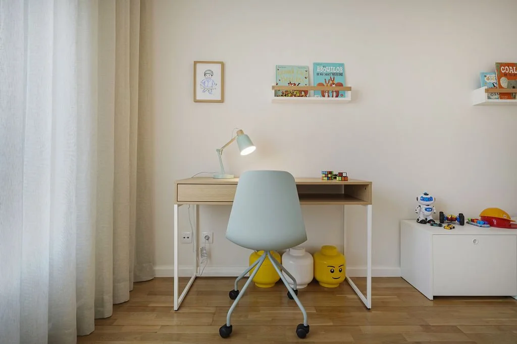 Escritório infantil com mesa de madeira clara, cadeira branca, abajur, estantes com livros e brinquedos, brinquedo robô, e vasos decorativos com rostos, em um ambiente com cortinas brancas e piso de madeira.