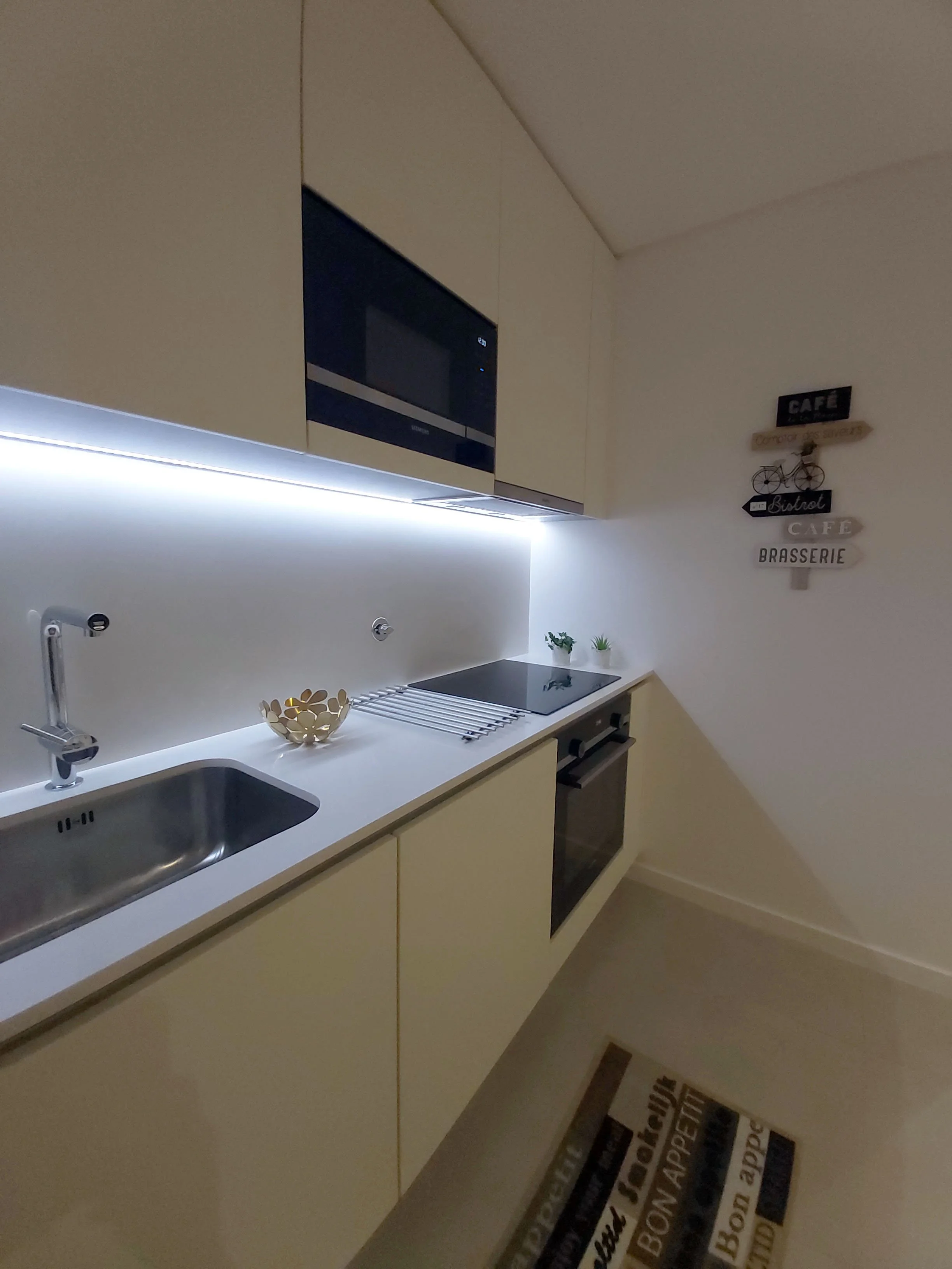 Home Staging de Apartamento em Quarteira, Algarve.
Cozinha moderna com armários brancos, micro-ondas embutido, cuba em aço inox, placa de indução, alguns pequenos vasos com plantas, decoração de parede com palavras relacionadas a café e bistro, tapet