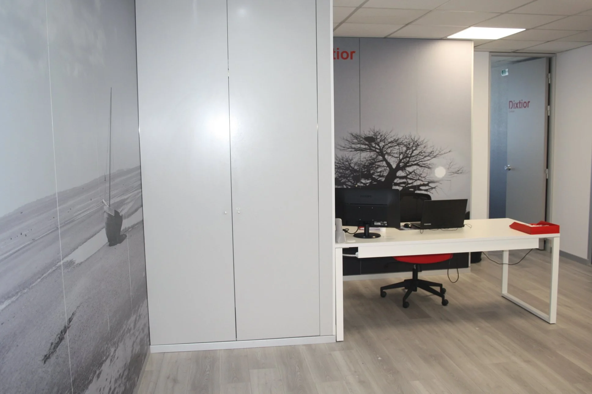 Escritório com parede decorada com uma árvore e uma paisagem de praia em tons de cinza, com mesa branca, computador, cadeira com encosto vermelho, e uma porta ao fundo.