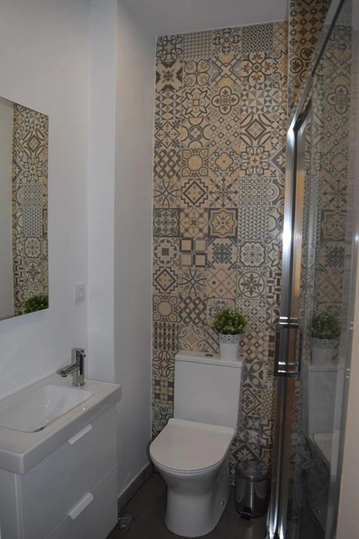 Banheiro com parede de azulejos decorativos, vaso sanitário, espelho, pia com armário, plantas decorativas, e box de vidro.