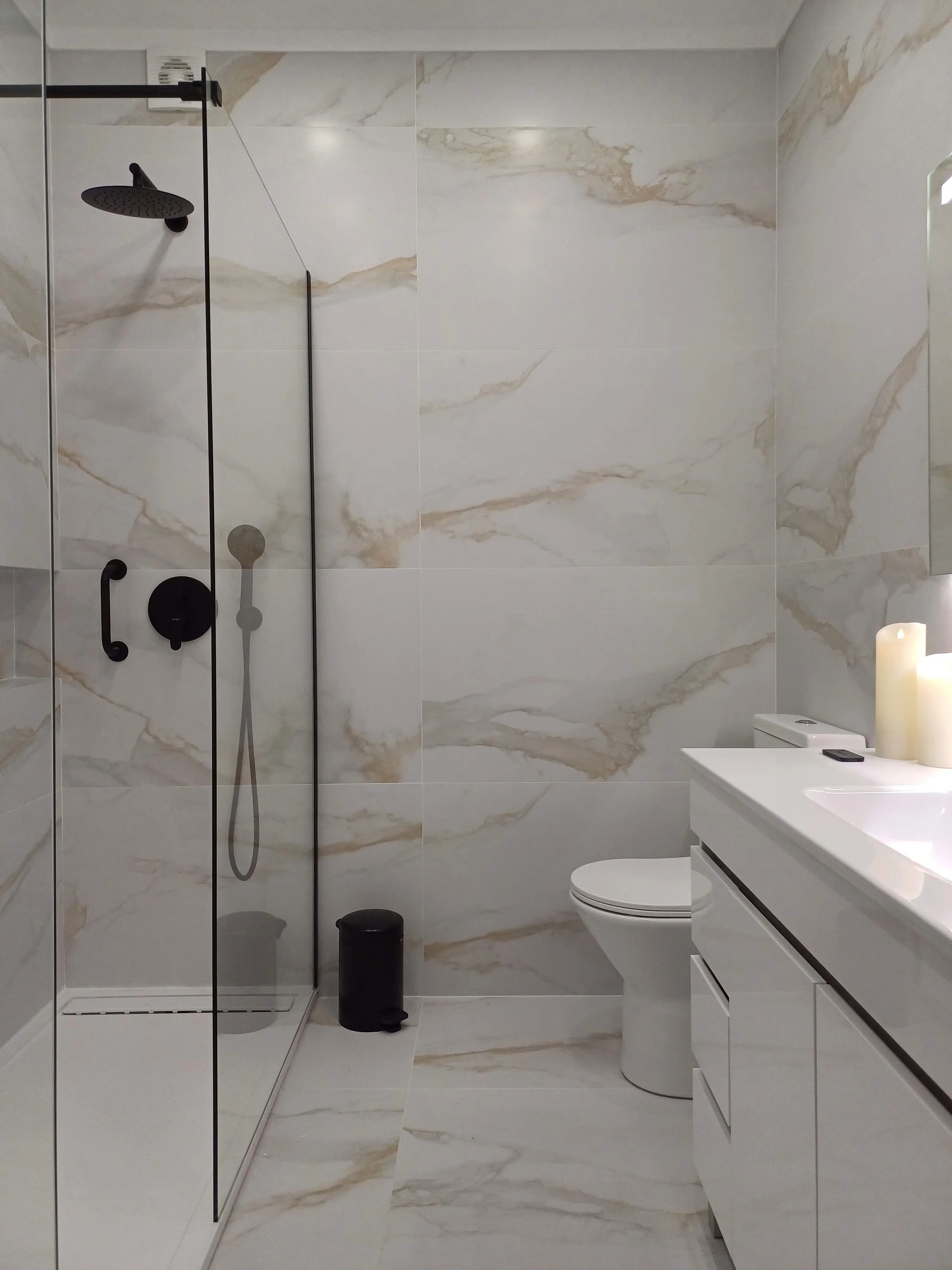 Banheiro moderno com parede e piso de mármore branco com veios dourados, ducha de vidro com torneira preta, pia branca com objetos de decoração e velas.