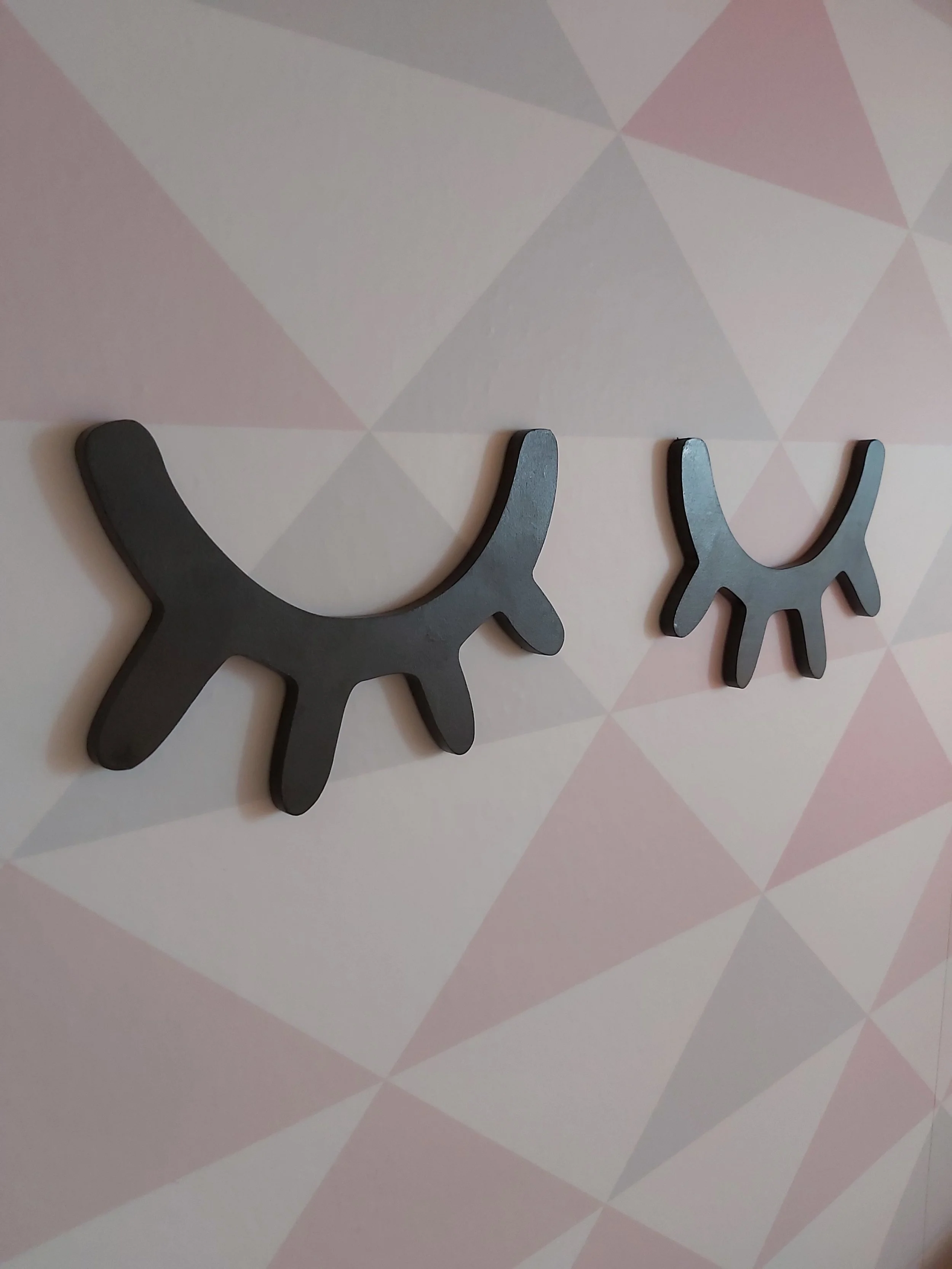 Duas molduras pretas com formato de cílios decorativos na parede com fundo de padrão geométrico em tons de rosa, cinza e branco.
