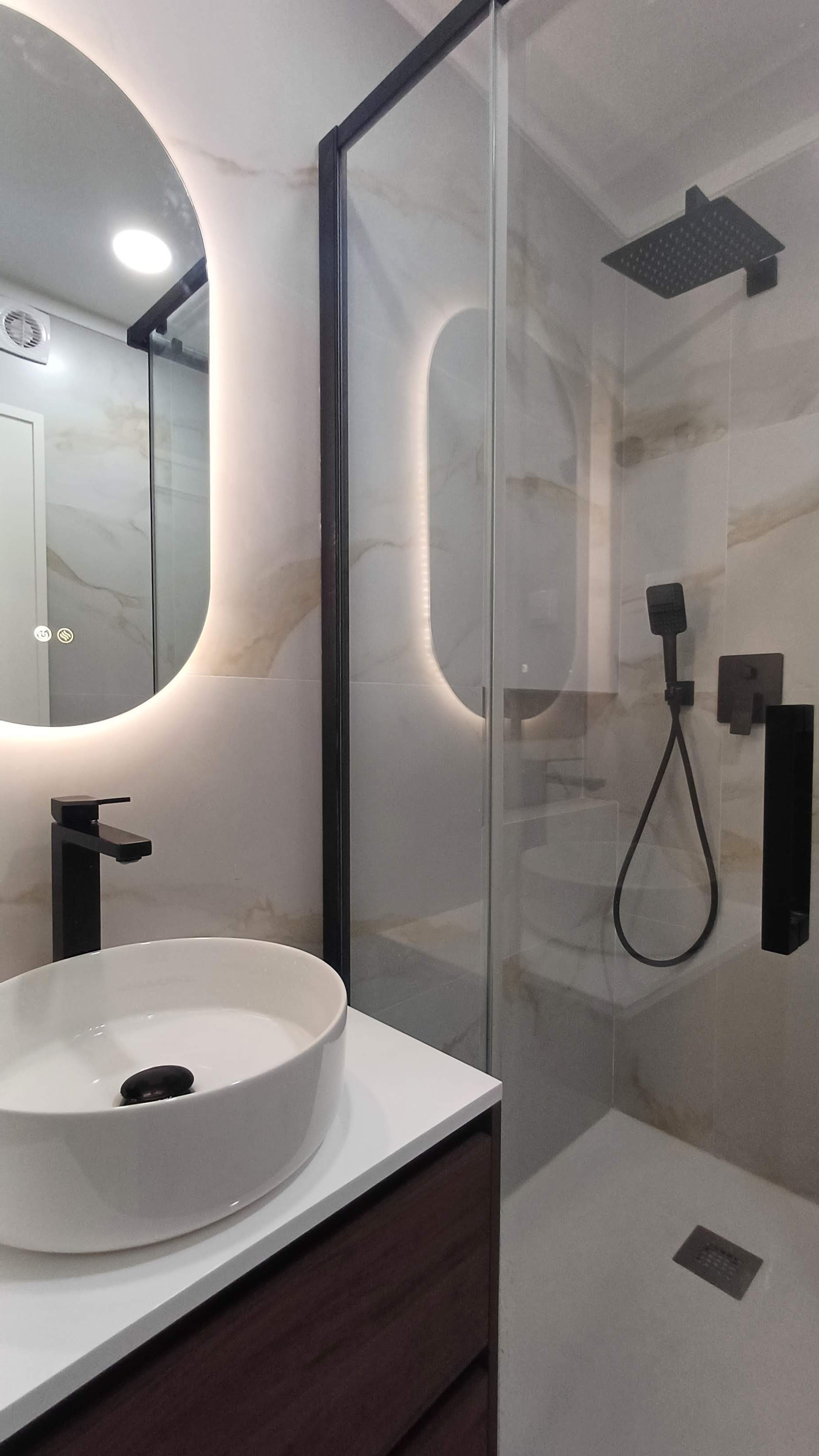 Banheiro com pia de cuba redonda branca, espelho com iluminação de LED ao redor, parede de mármore claro, cabine de ducha com porta de vidro, ducha de teto preta, e acessórios de banheiro em preto.