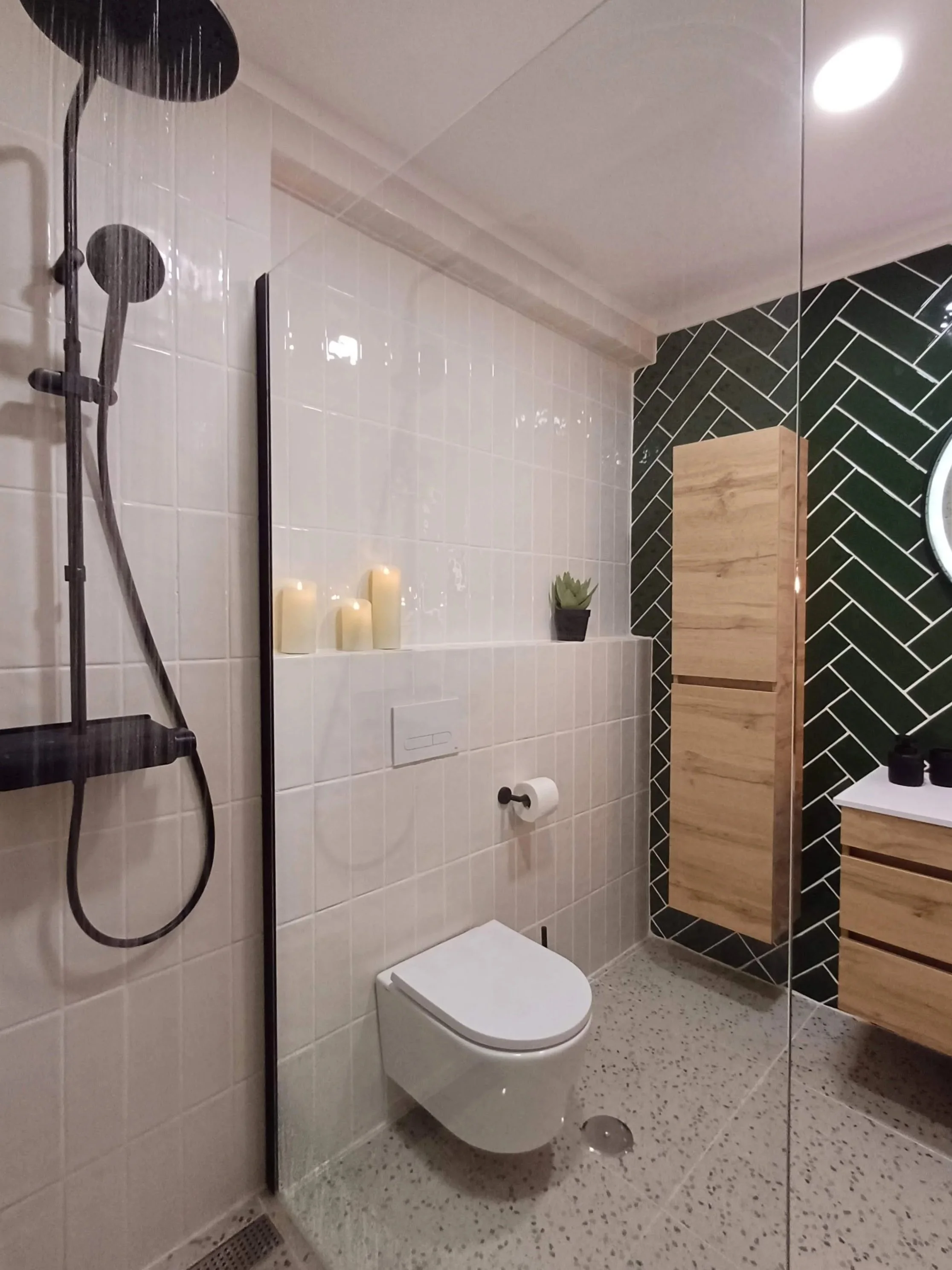 Banheiro moderno com box de vidro com ducha preta, vaso sanitário branco, prateleira com velas e planta, parede com azulejos brancos, armário de madeira e parede com azulejos pretos em padrão diagonal.