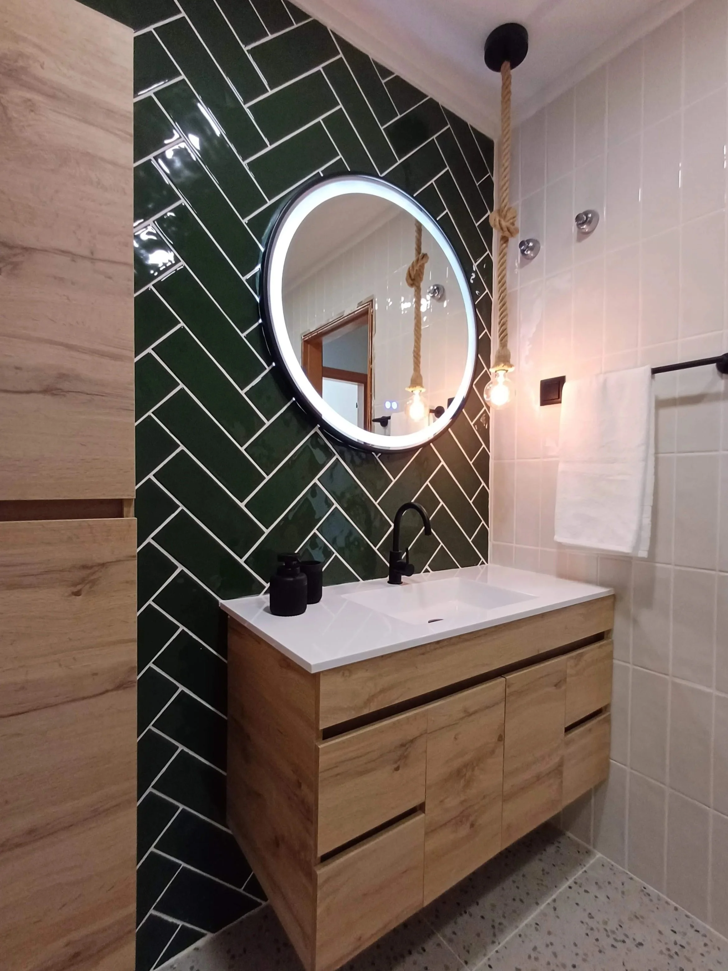 Espelho redondo com luz de LED sobre pia de pia de madeira com vaso e saboneteiro preto, parede com azulejos verde escuro em padrão de espinha de peixe, parede ao lado com azulejos brancos, luminárias com corda pendurada no teto.