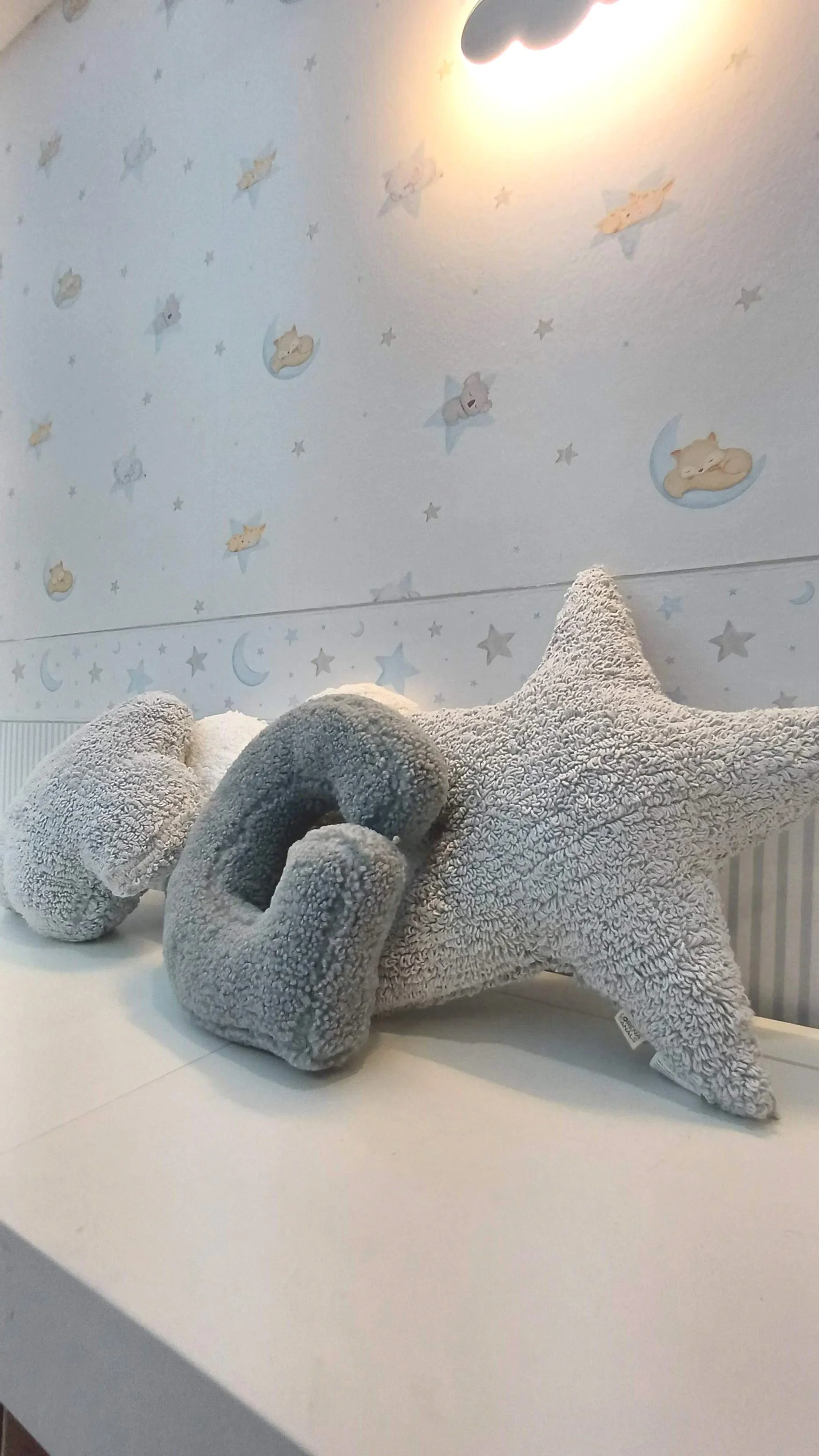 Imagem de dois bonecos de pelúcia, um cinza escuro e outro cinza claro, sobre um trocador branco, com um fundo de parede decorada com desenhos de ursinhos, estrelas e luas em tons suaves.