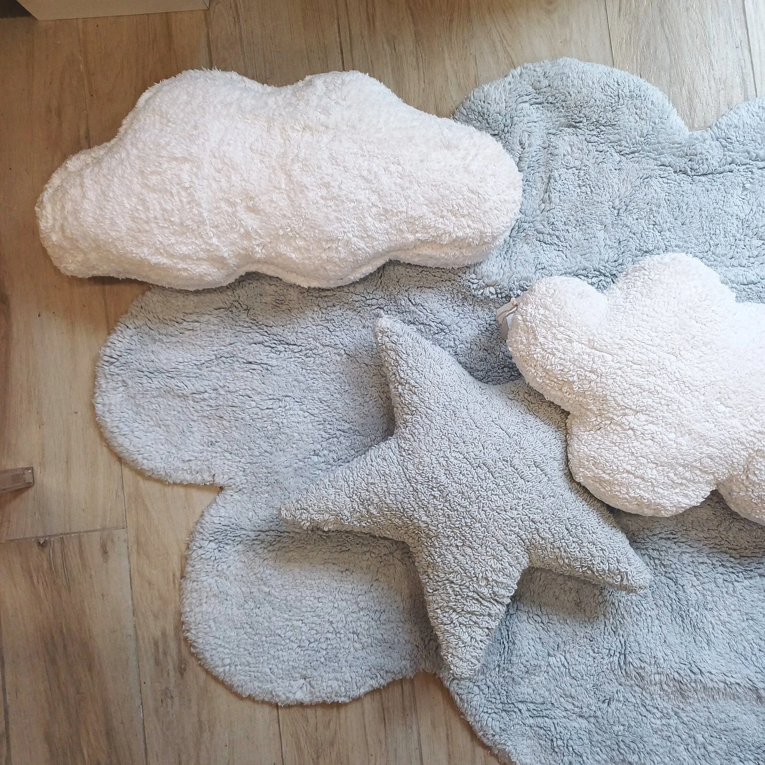 Conjunto de tapetes e almofadas em formato de nuvem e estrela, feitos de tecido felpudo, em cores branco, cinza claro e azul claro, dispostos sobre piso de madeira.