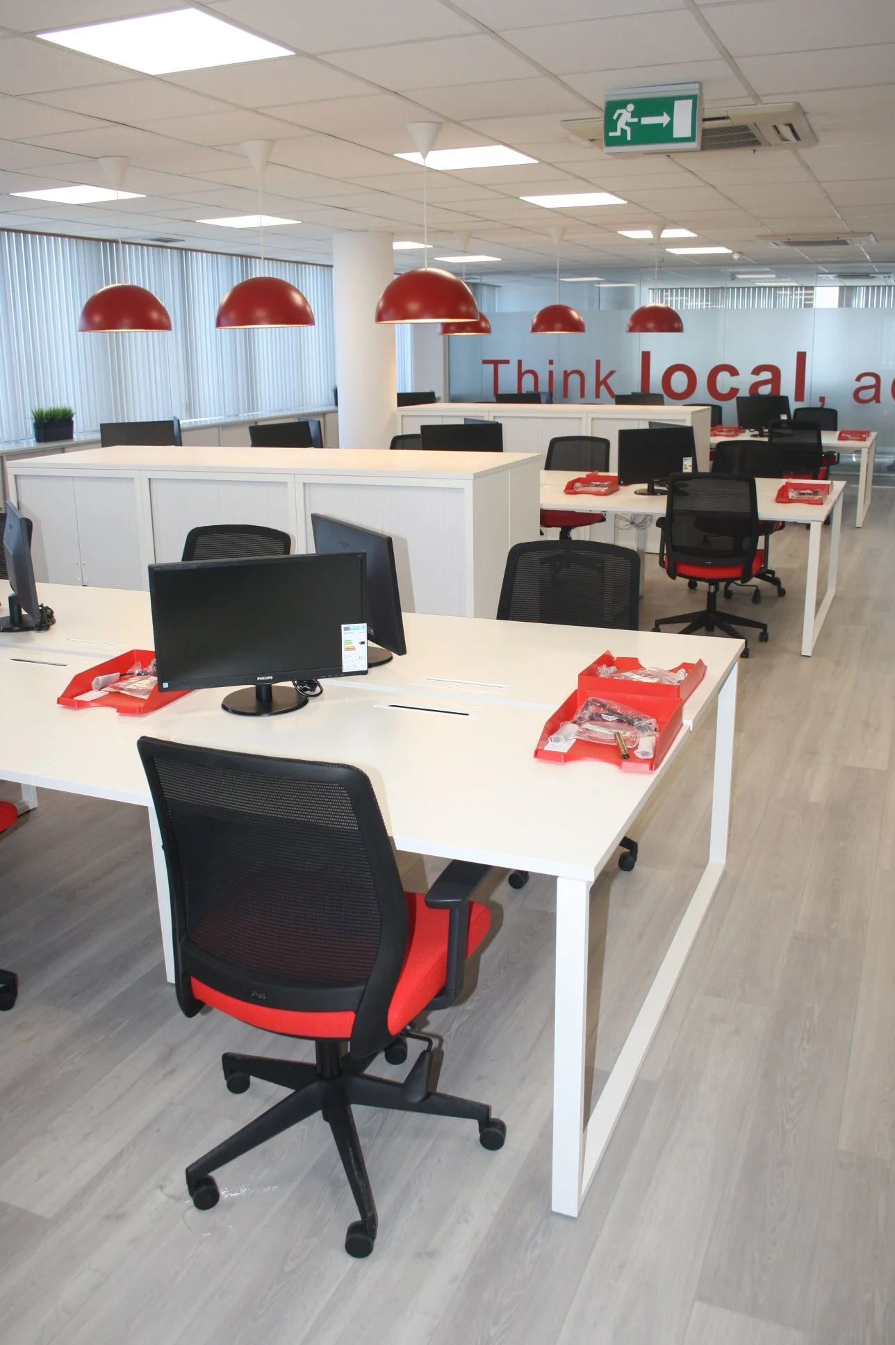 espaço de escritório com mesas de trabalho e cadeiras pretas e vermelhas, computadores, pranchetas vermelhas, iluminação de teto e parede de vidro com uma frase "Think local"