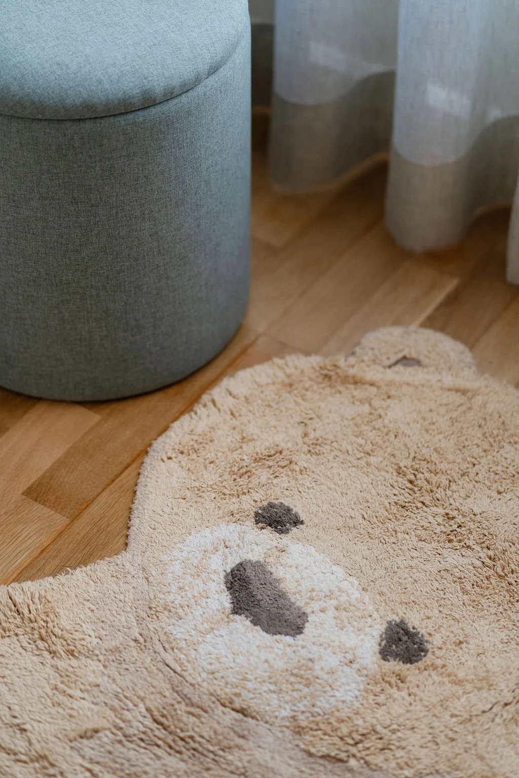 Tapete de pelos longos bege com manchas pretas e brancas, colocado ao lado de um móvel cinza e cortinas de tecido claro, em piso de madeira.