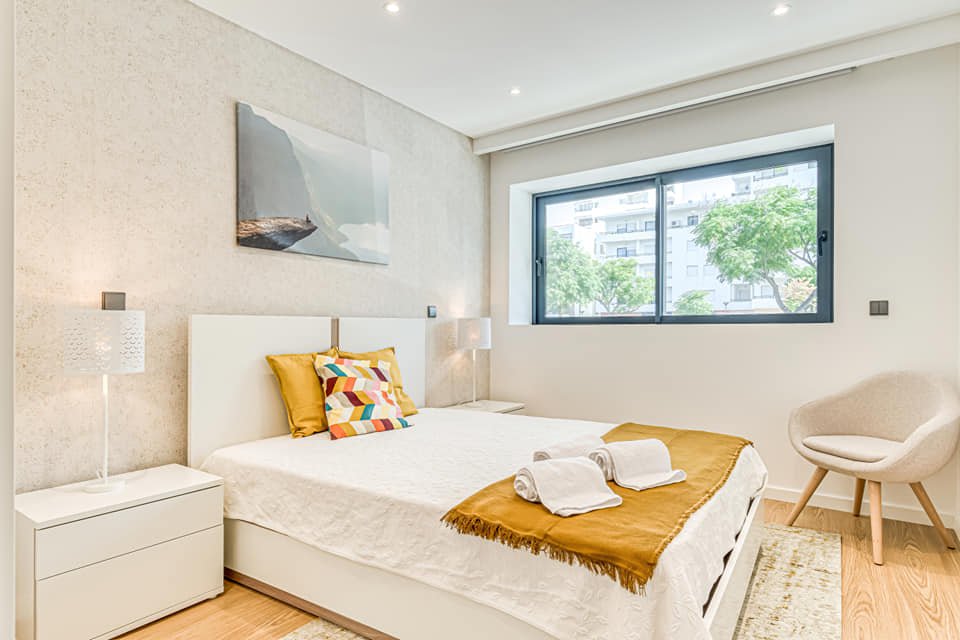 Quarto bem iluminado com cama de casal, travesseiros coloridos, janelas grandes com vista para árvores e prédios, cômodo decorado em tons neutros, com mesa de cabeceira e poltrona confortável ao lado.