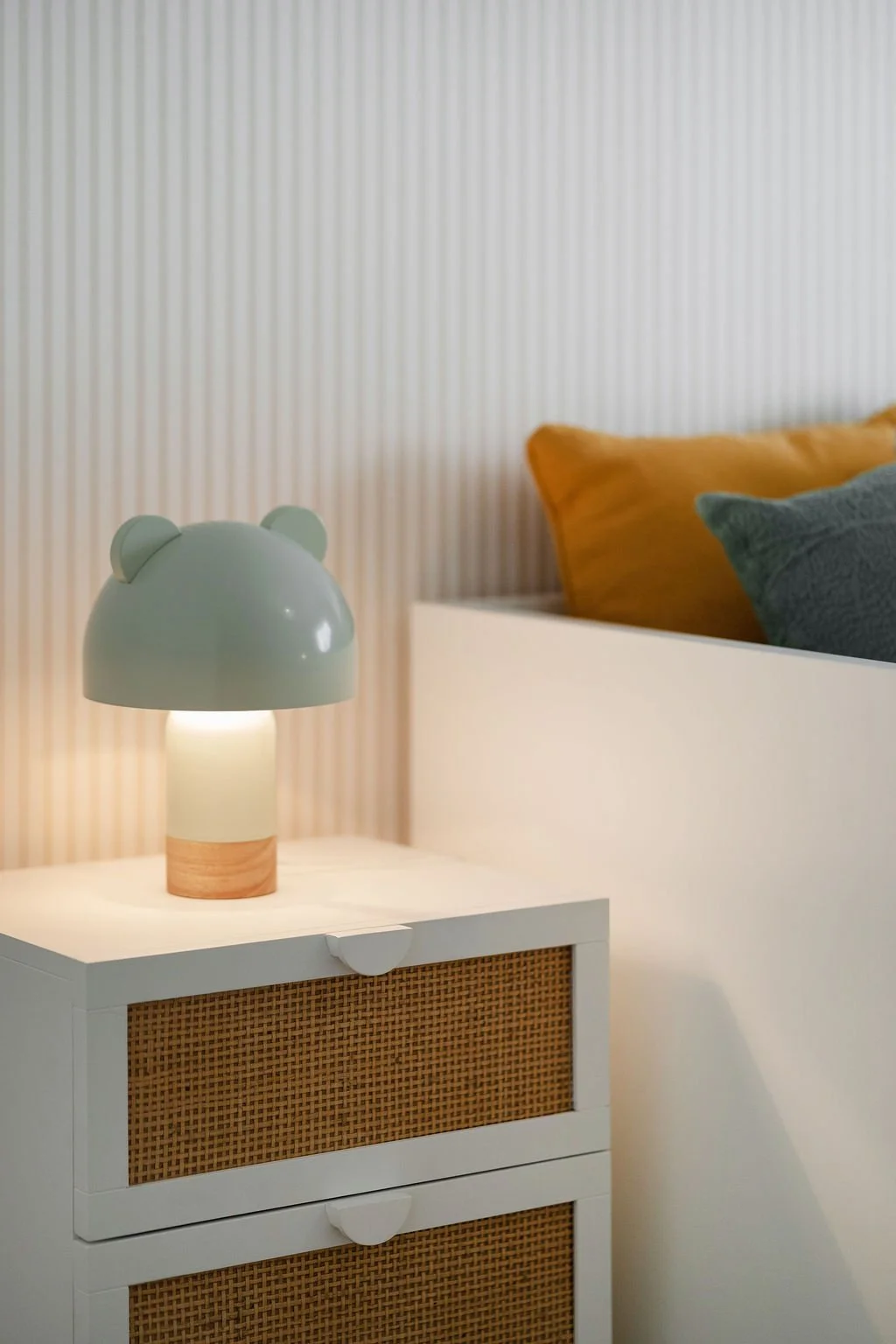 Abajur com possibilidade de design de rosto de urso, em cores pastel, sobre criado-mudo branco com portas de fibra de vime, ao lado de cama com travesseiros amarelos e cinzas, em quarto com parede de papel de parede listrado.