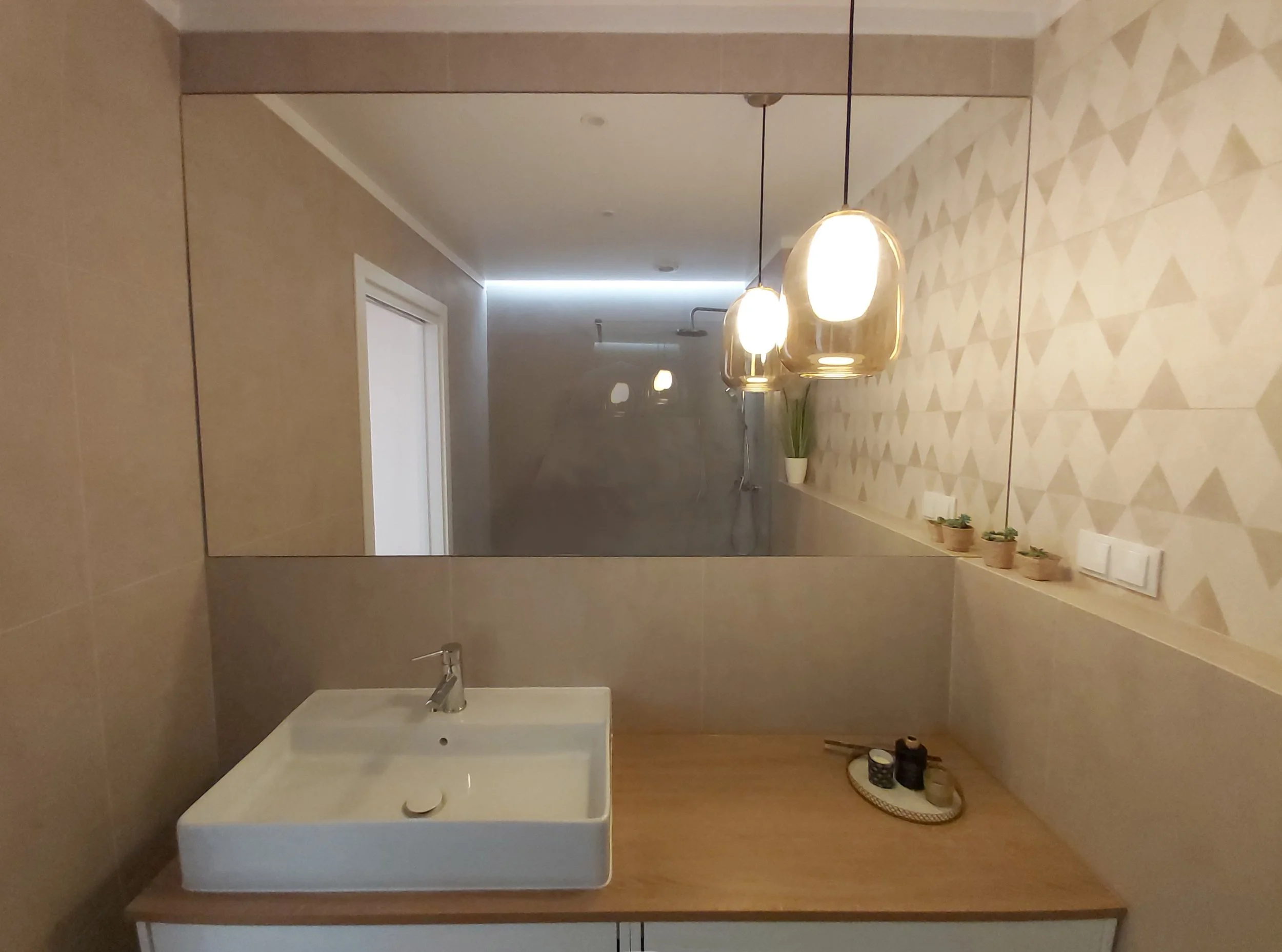 Espelho grande refletindo uma parede com iluminação de lâmpadas suspensas, bancada de madeira com uma cuba branca, pequenas plantas em vasinhos, pentes, velas e utensílios de cosmética.