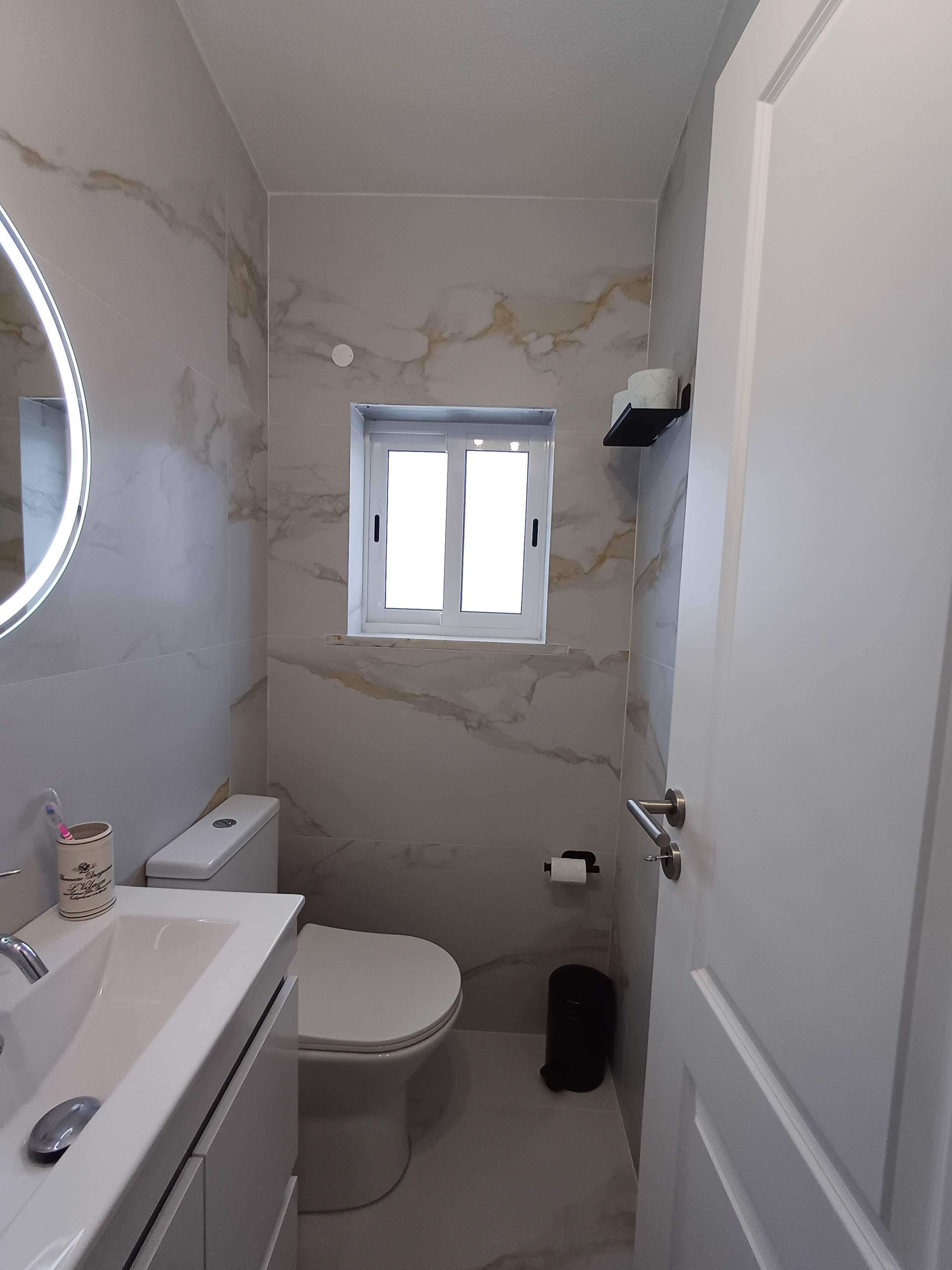 Banheiro com paredes de mármore bege com veios dourados, janela no centro da parede do fundo, vaso sanitário, pia branca com espelho de luz ao lado, e itens de higiene no balcão.