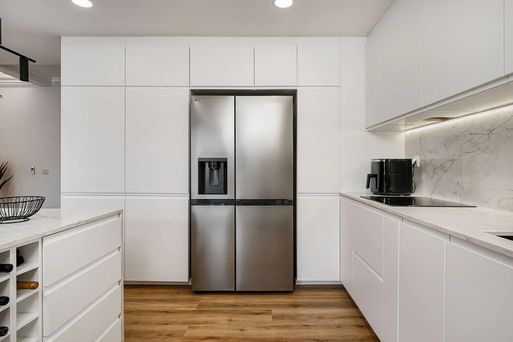 Cozinha moderna com armários brancos e ante panele de mármore, geladeira de aço inoxidável na parede ao centro, e eletrodomésticos pretos, incluindo cafeteira elétrica, ao longo da bancada à direita.