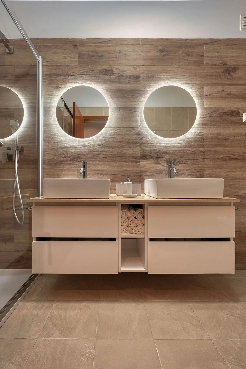 Decoração Casa-de-Banho. Revestimento cerâmico imitação madeira, Piso em cerâmica, Armário duplo suspenso branco lacado com 2 espelhos de parede redondos com iluminação integrada.