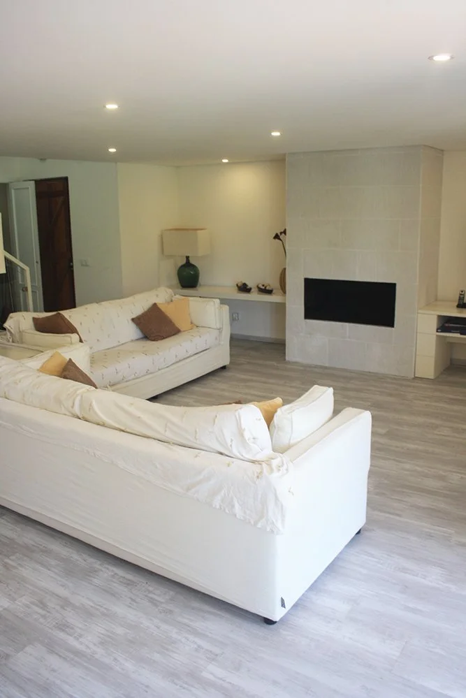 Sala de estar com sofá branco, almofadas em cores neutras, lareira de parede de pedra clara, luminárias de teto embutidas, vaso com plantas e luminária de mesa ao fundo.