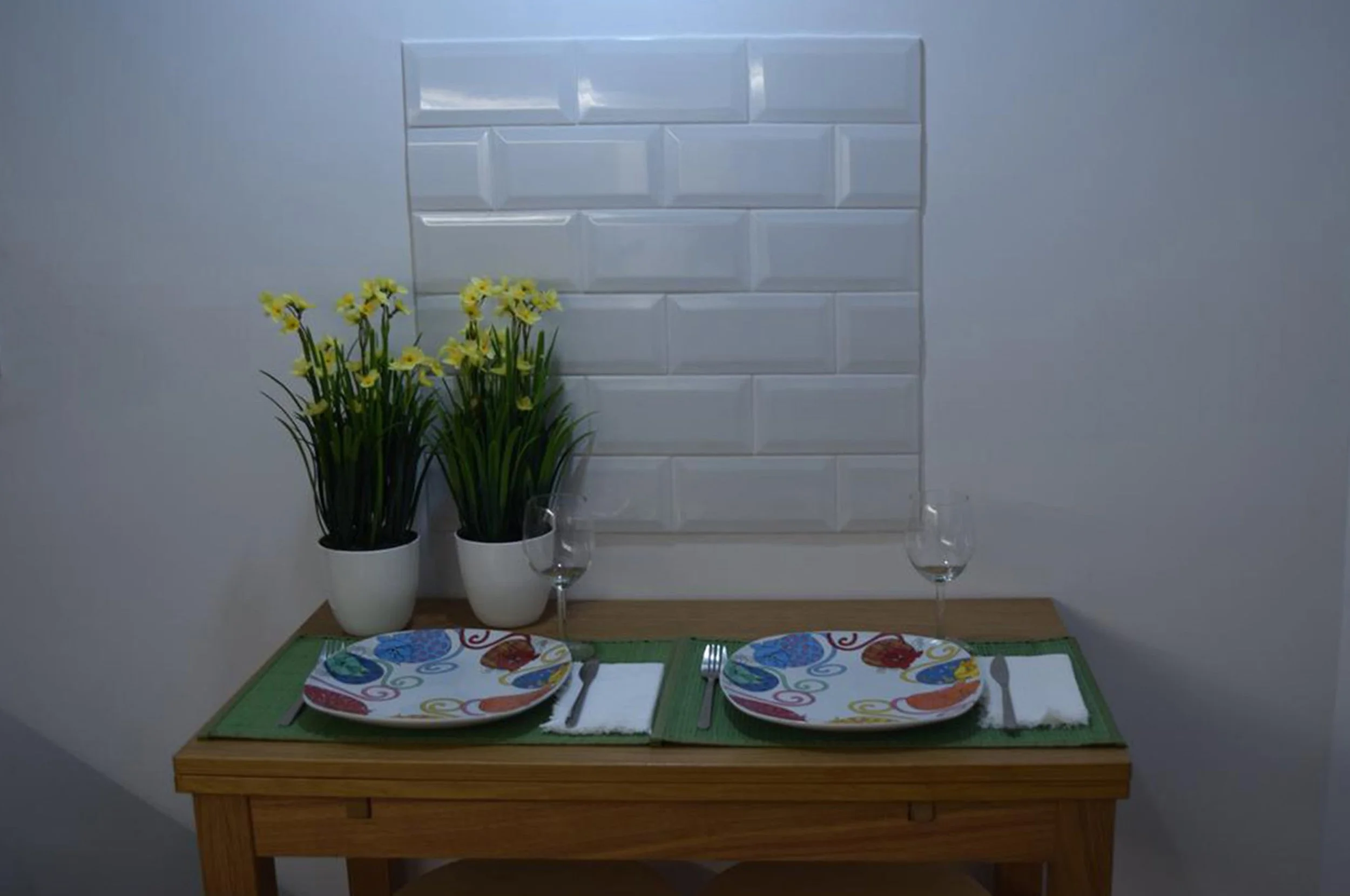 Mesa de jantar pequena com duas louças decoradas, talheres e copos vazios, duas plantas com flores amarelas ao fundo e um revestimento de azulejos brancos na parede.