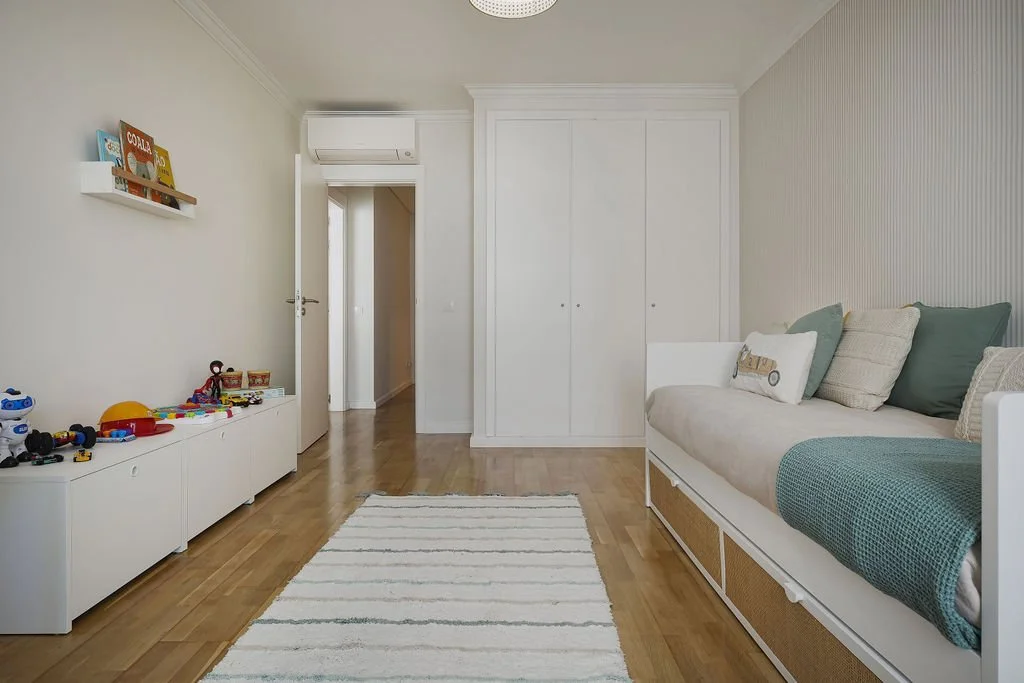 Sala de estar com piso de madeira, sofá branco com almofadas, brinquedos infantis em móvel branco, prateleira com livros, armário branco e cortina branca na janela.