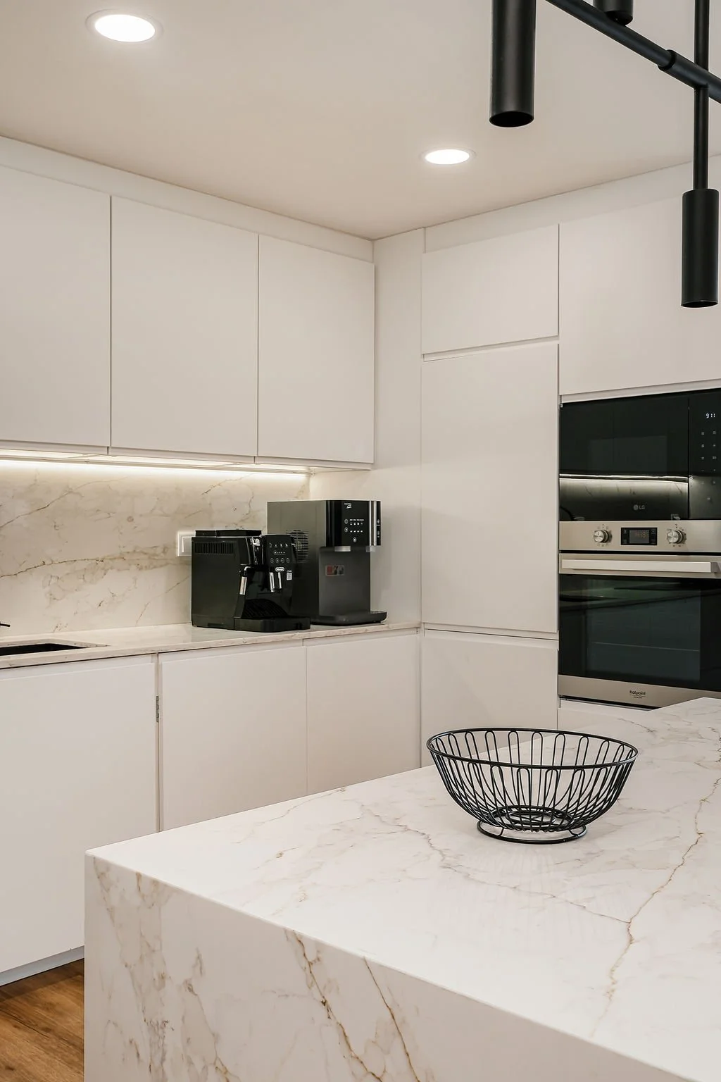 Cozinha moderna com bancadas de mármore branco, armários brancos e eletrodomésticos de aço inox, incluindo uma máquina de café expresso, uma cafeteira e um forno embutido. Há uma ilha central com uma cesta de metal preta.