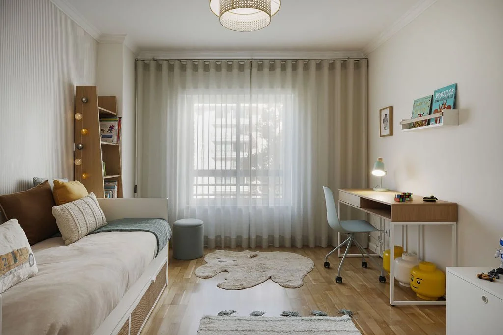 Quarto bem iluminado com sofá de cama branco, almofadas coloridas, estante de madeira com luzes decorativas, janela grande com cortinas brancas, área de estudo com mesa de madeira, cadeira cinza, luminária, prateleira com livros, tapete em formato de