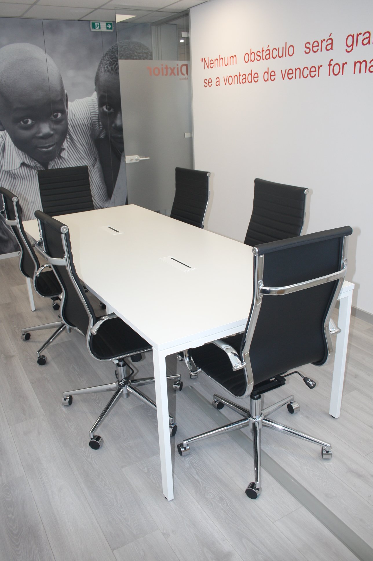 Sala de reunião com mesa branca e seis cadeiras pretas com estrutura de metal prateada, parede com foto de duas crianças negras e citação na parede em português.