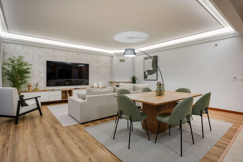 Decoração de Sala com área de estar e área de jantar. Aplicação de papel de parede, tapetes e acessórios de decoração. Candeeiro de pé e sanca com iluminação indirecta. Poltrona, mobiliário de TV e plantas