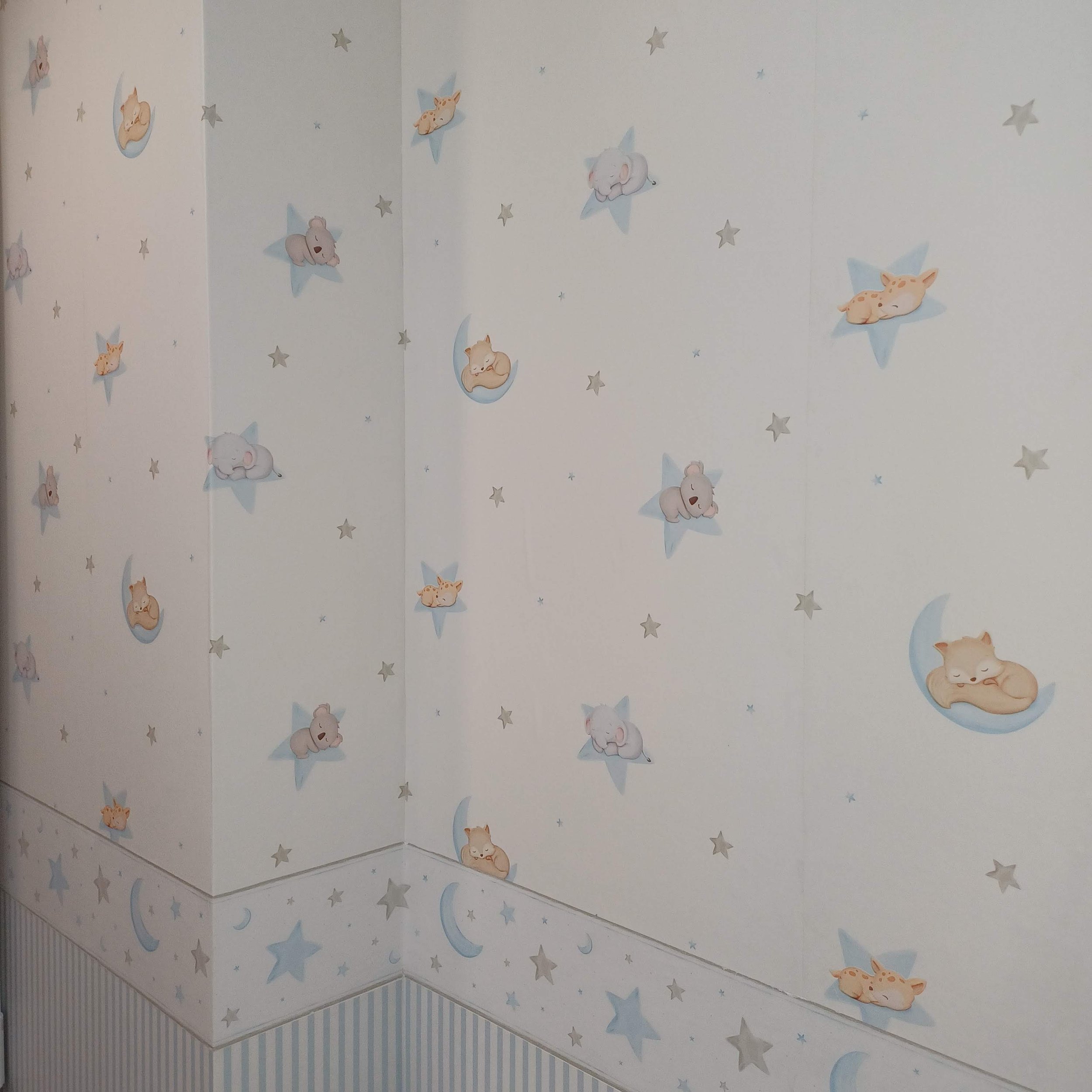 Parede com papel de parede de fundos branco e desenhos de animais de dormir em estrelas e luas, incluindo gatinhos e coelhinhos em tons suaves de bege, cinza, azul claro e branco.