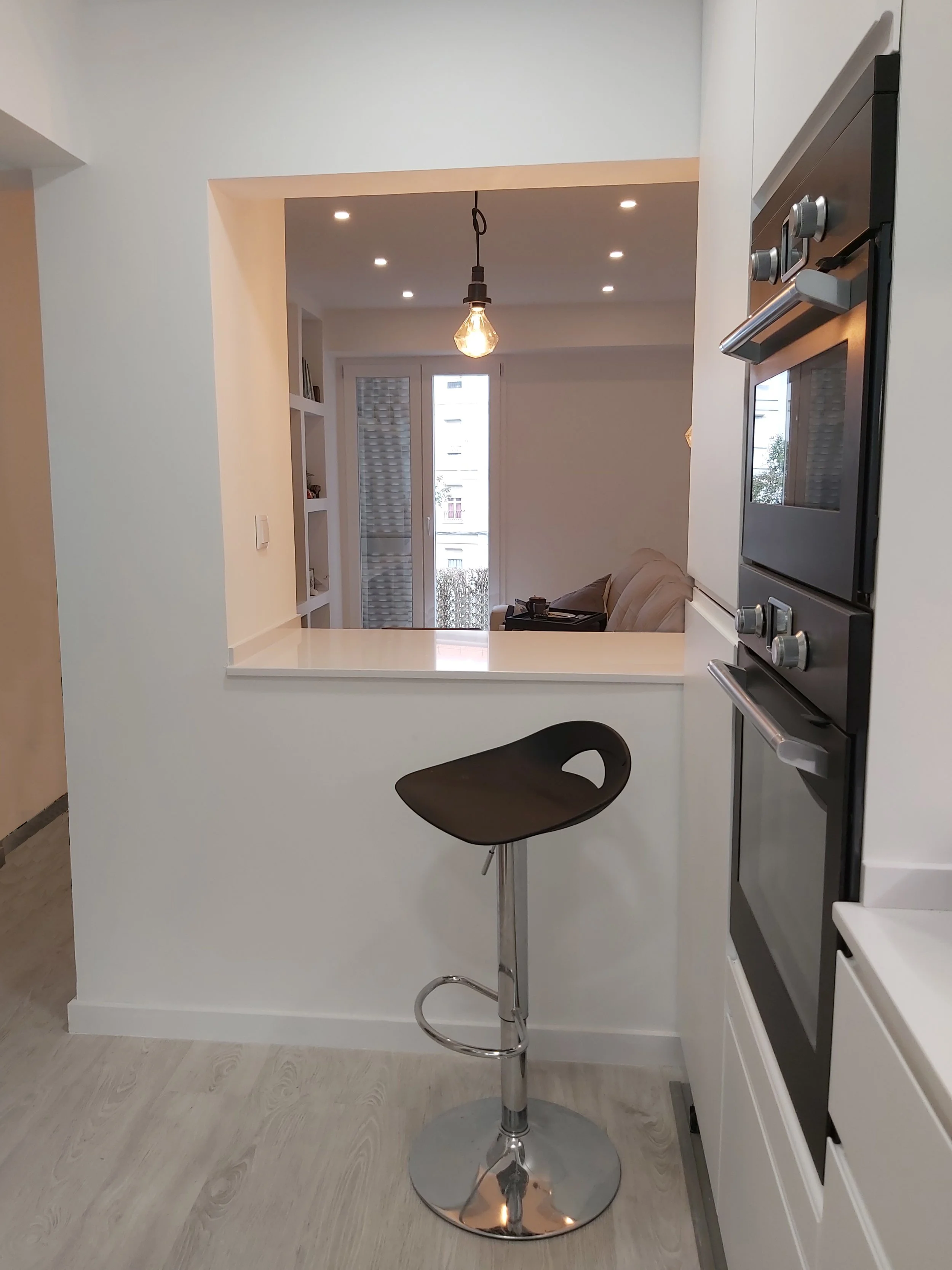 Barra de café com assento giratório preto em cozinha moderna com iluminação embutida e sala de estar ao fundo.