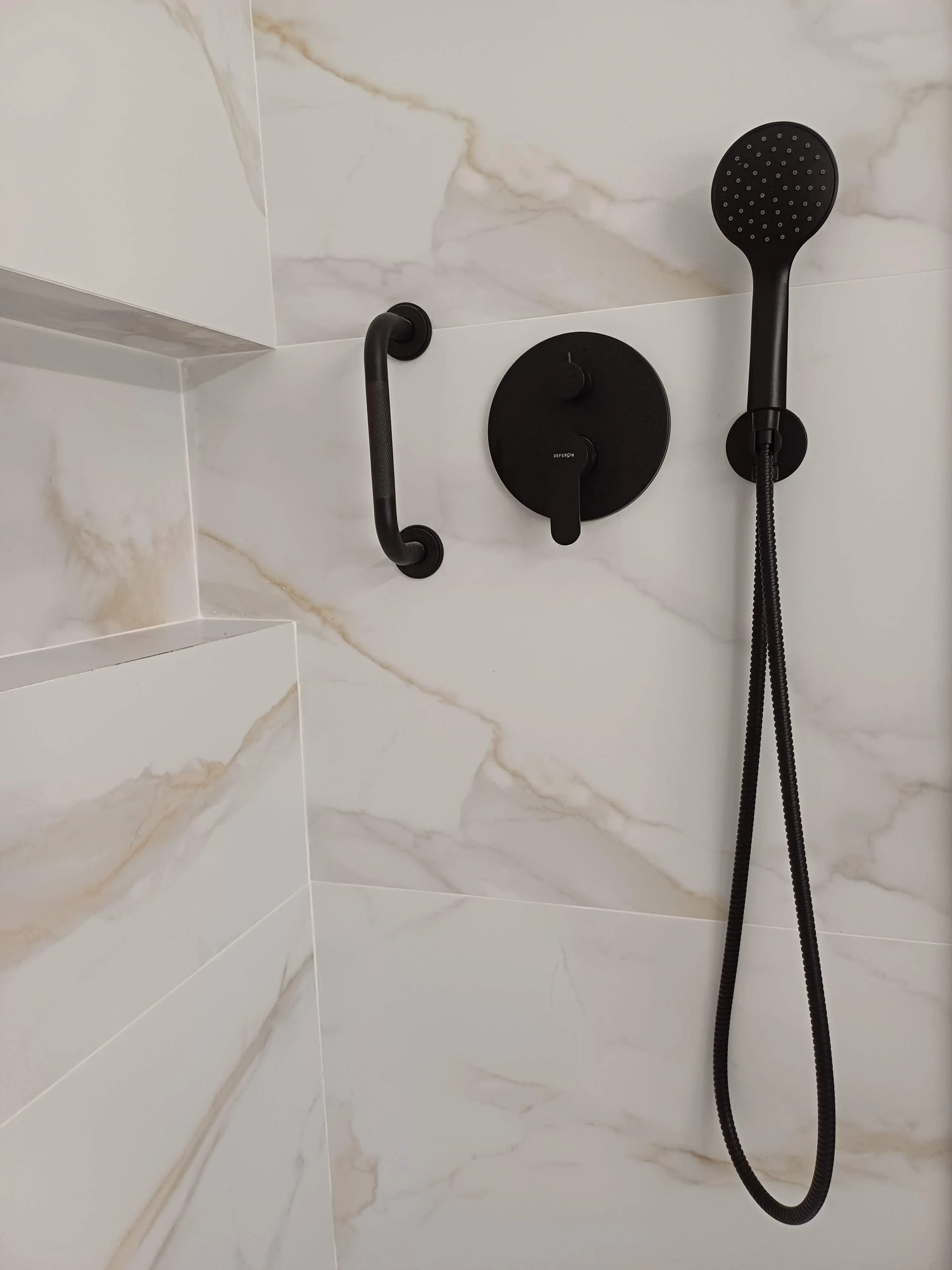 Chuveiro preto com controle de temperatura no banheiro, com uma barra de apoio ao lado, parede de mármore branco com veios bege.