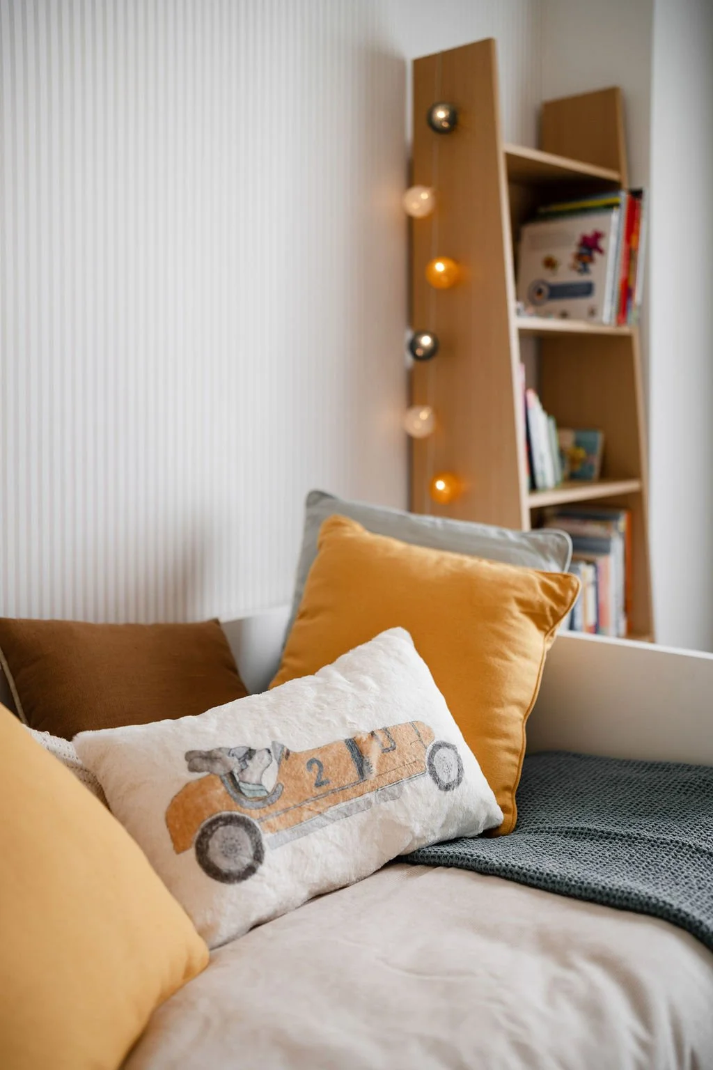 Cama com almofadas de cores bege, branca, cinza e marrom, ao lado de uma parede com prateleira de livros e uma iluminação decorativa com luzes de cores quente e fria.