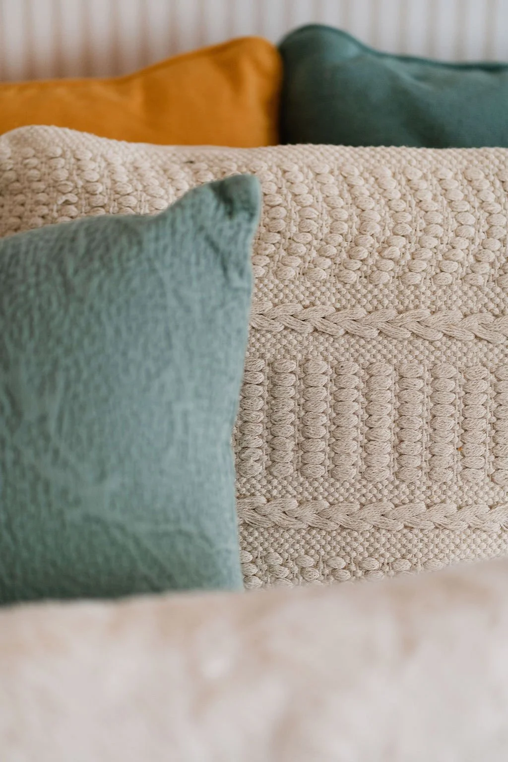 Diversos travesseiros de tecido com diferentes texturas e cores, em tons neutros, branco, verde e marrom, dispostos em um sofá ou cama.