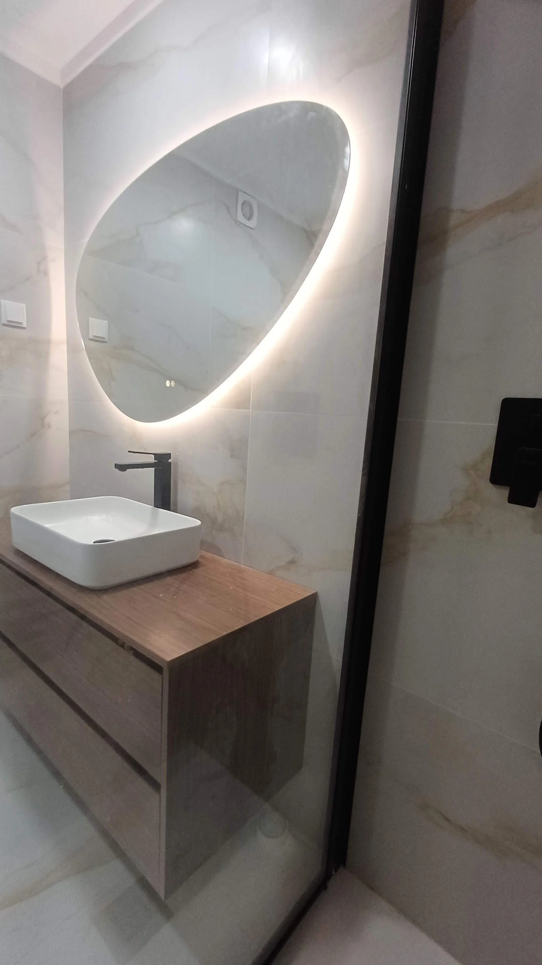 Espelho oval com iluminação LED ao lado de uma pia de cuba quadrada branca sobre uma bancada de madeira, em um banheiro com paredes de mármore claro.