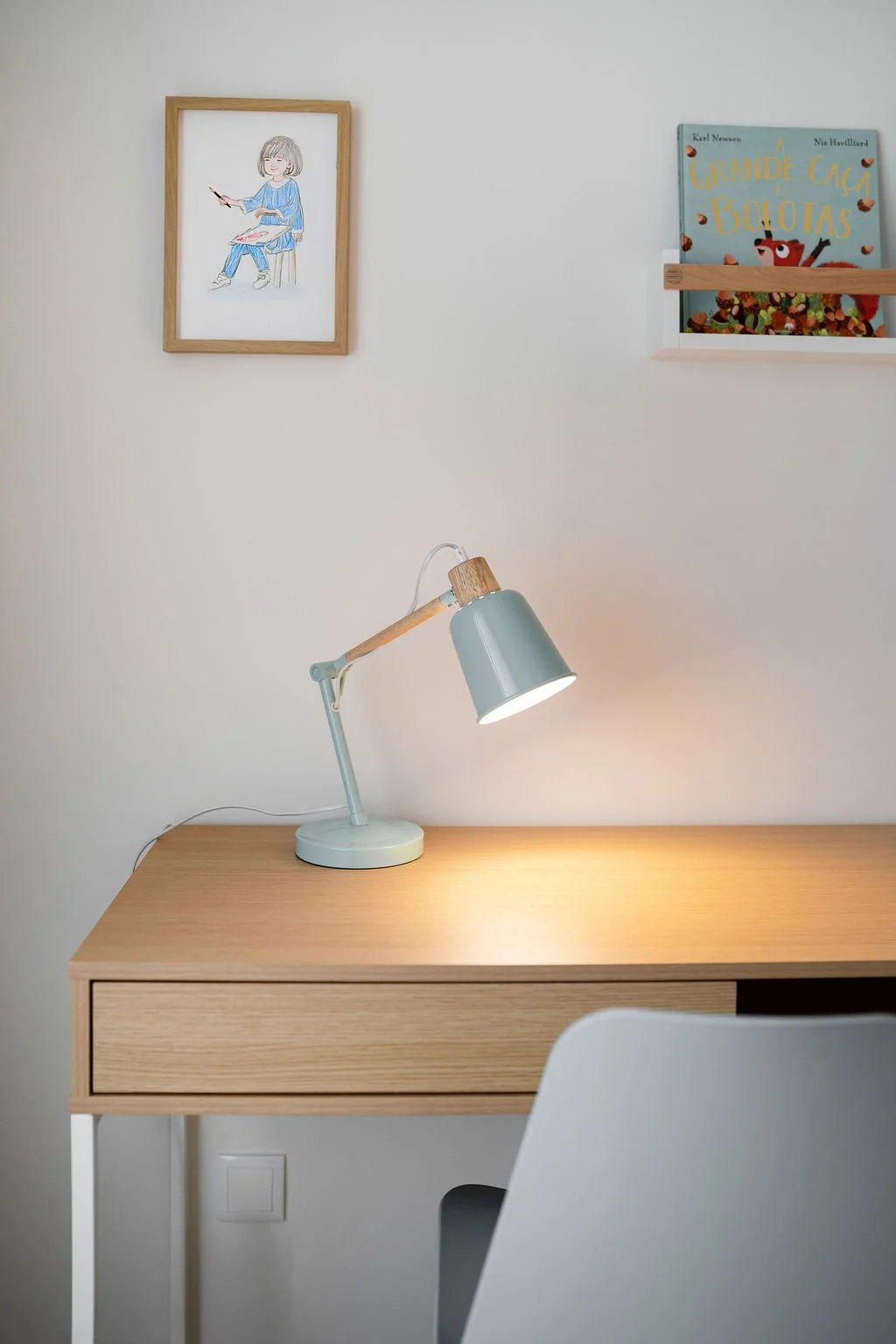 Mesa de escritório de madeira clara com luminária de mesa azul acesa, parede branca com quadro de desenho de menina pintando, e uma estante com livro infantil ao lado.