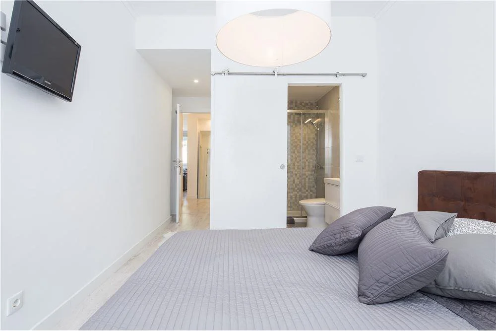 Quarto com cama de roupa cinza e três travesseiros cinza, parede branca, televisão na parede e banheiro visível ao fundo.