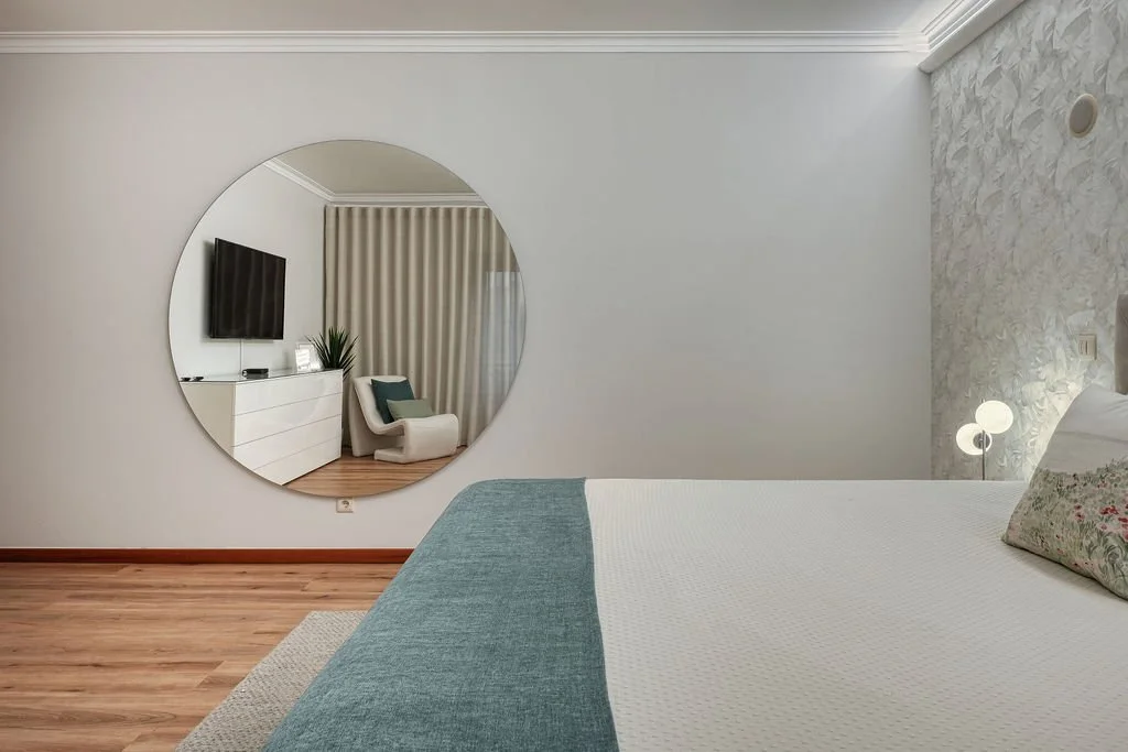 Quarto com cama, televisão na parede, espelho redondo grande, poltrona, cortinas e luminária de mesa.