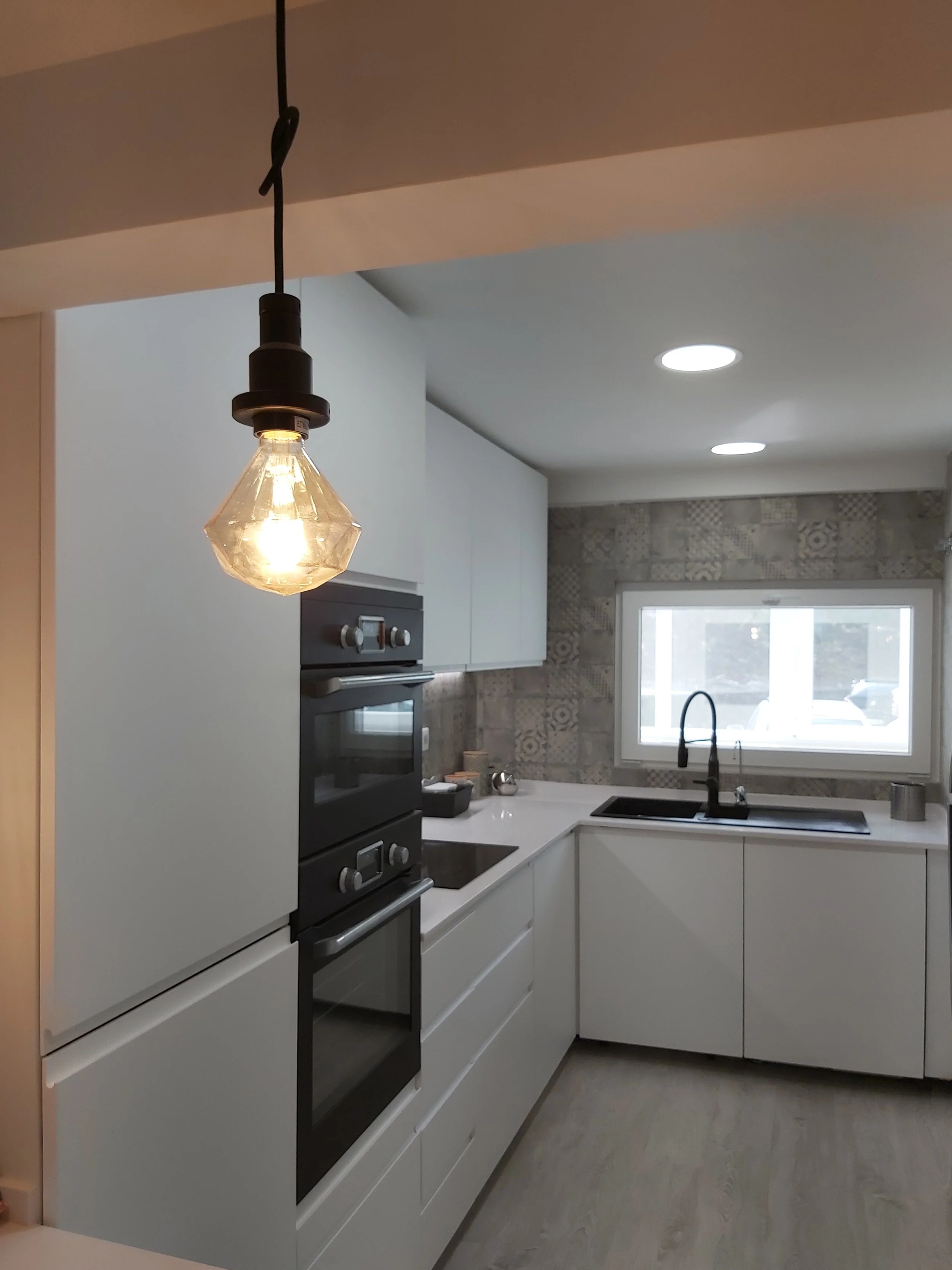 Cozinha moderna com armários brancos, forno embutido, pia com torneira preta, parede decorada com azulejos em tom cinza, janela grande e iluminação de teto.