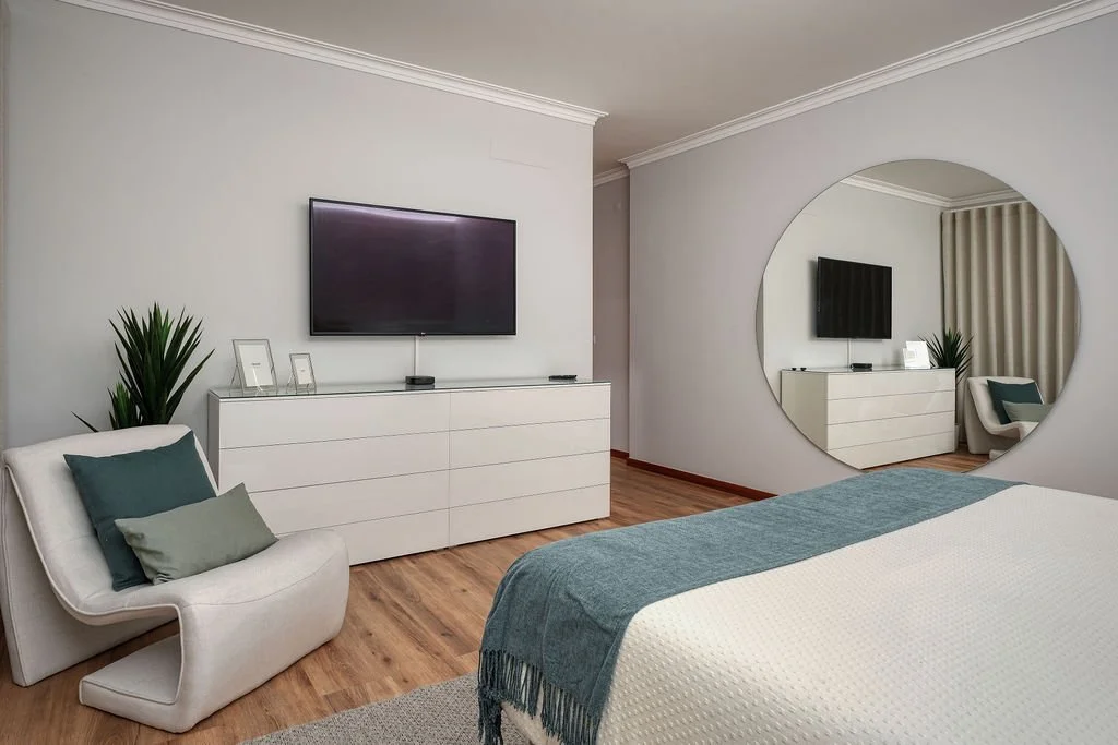 Quarto com parede branca, televisão na parede, com uma cômoda branca abaixo, decorada com quadros, planta verde, espelho redondo grande, cama com colcha branca e xale azul, cadeira branca com almofadas, e cortinas bege no fundo.