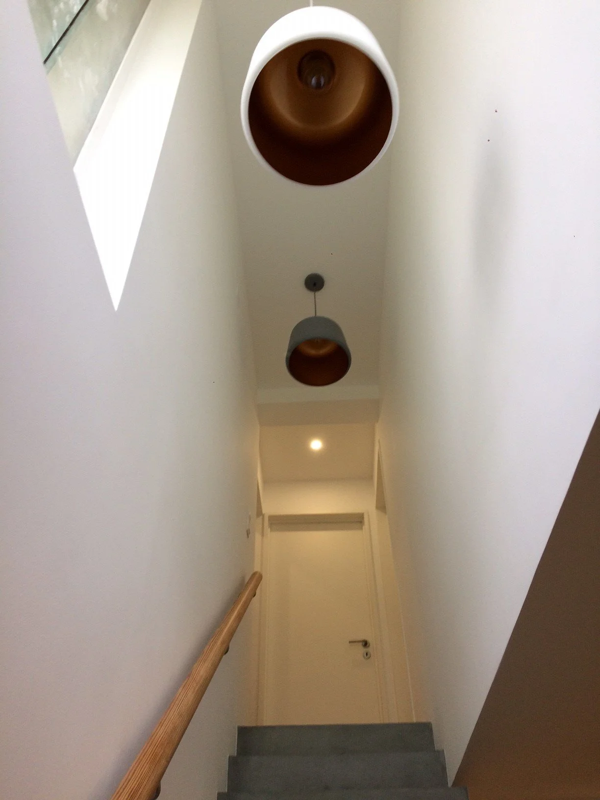 Escada de interior com corrimão de madeira, vista de baixo para cima, teto com duas luminárias pendentes e uma janela.