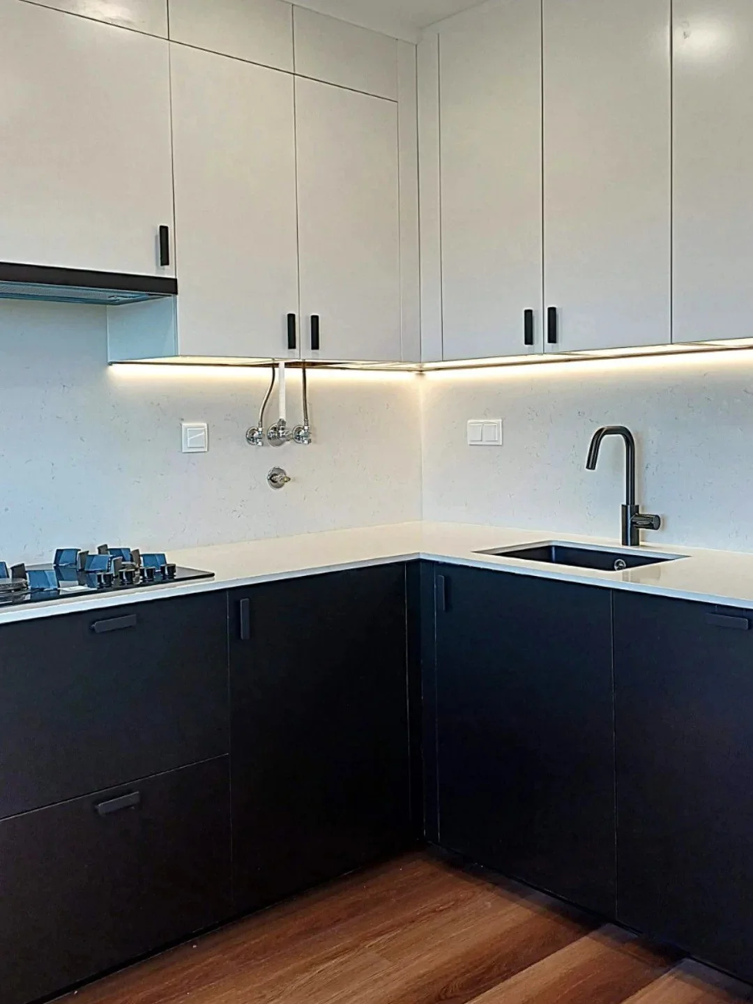 Cozinha moderna com armários brancos e pretos, pia preta, torneira preta, cooktop de gás, paredes claras e piso de madeira.