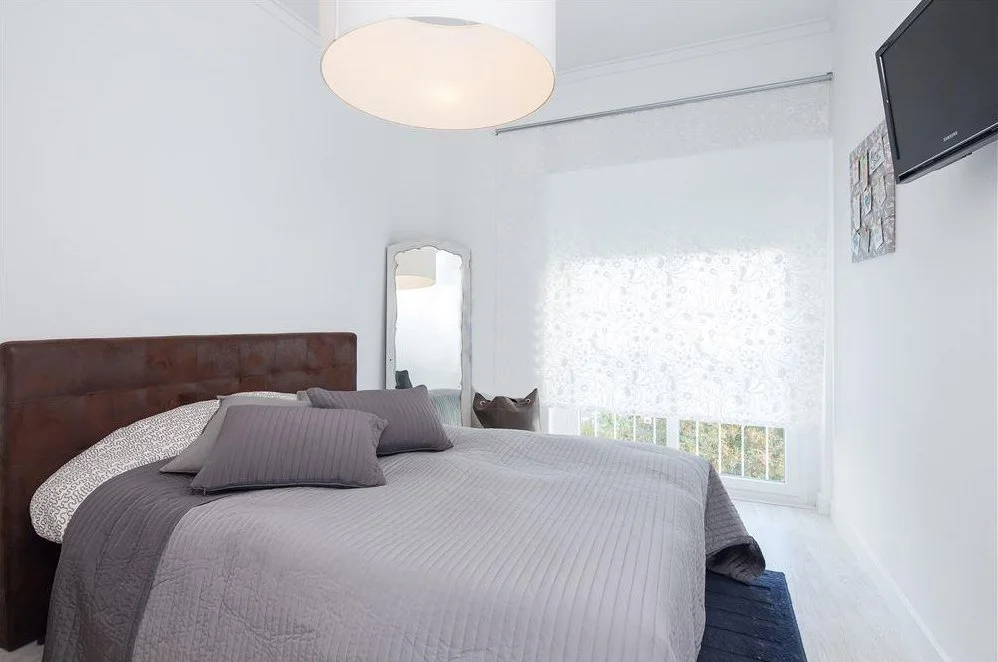 Quarto com cama de madeira, roupa de cama cinza e almofadas cinzas, espelho de corpo inteiro ao lado, janela com cortina branca, televisão na parede e luminária de teto